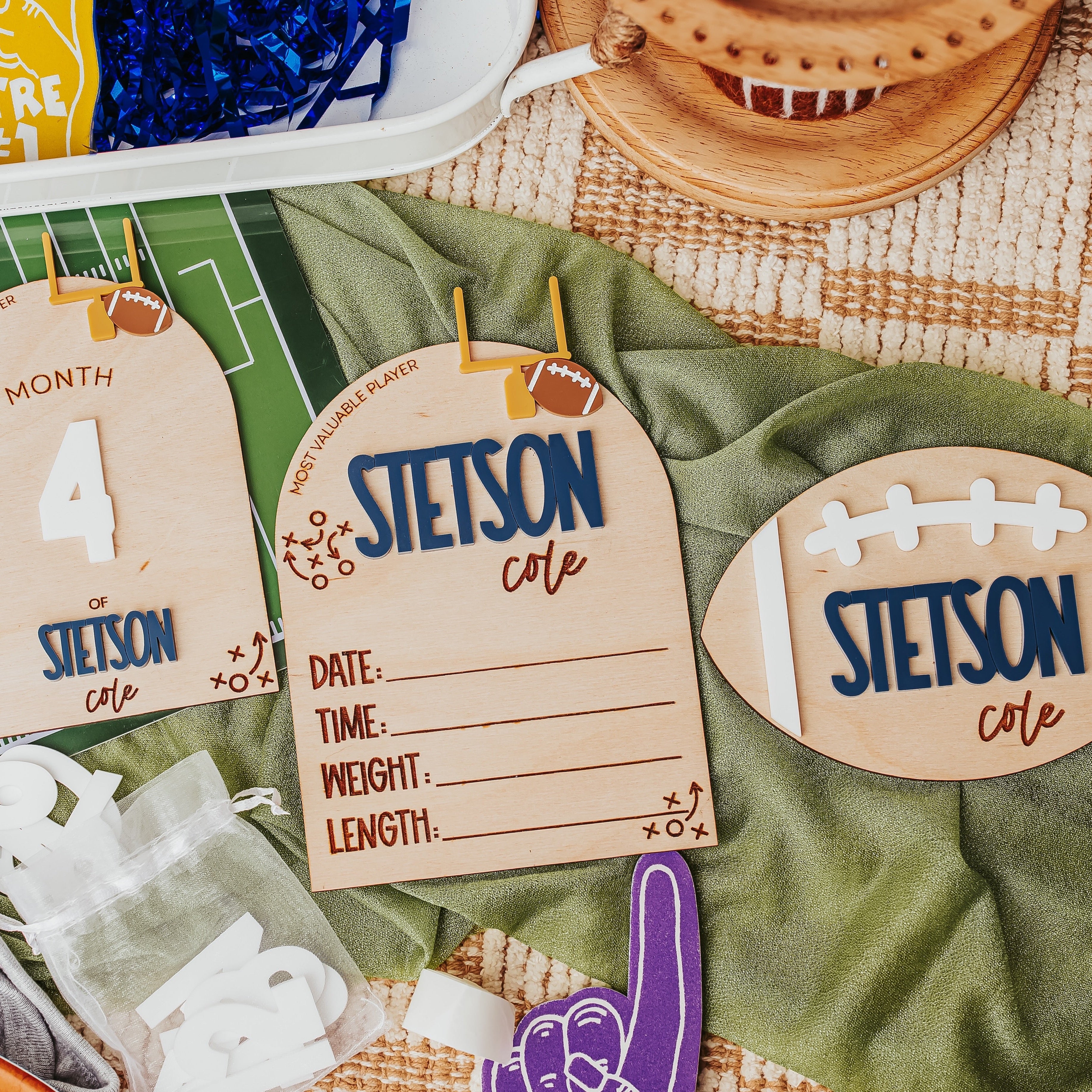 Custom football-themed baby milestone cards laid out on a green blanket with sports props, including a football-shaped card, a month card, and a birth details card.