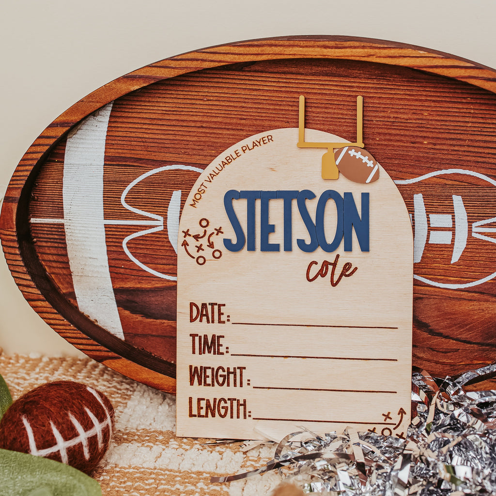 A wooden football-themed photo frame with a personalized name card reading 'STETSON cole' and sports fields printed on the wood, styled with a football, green fabric, and silver tinsel on a neutral background.