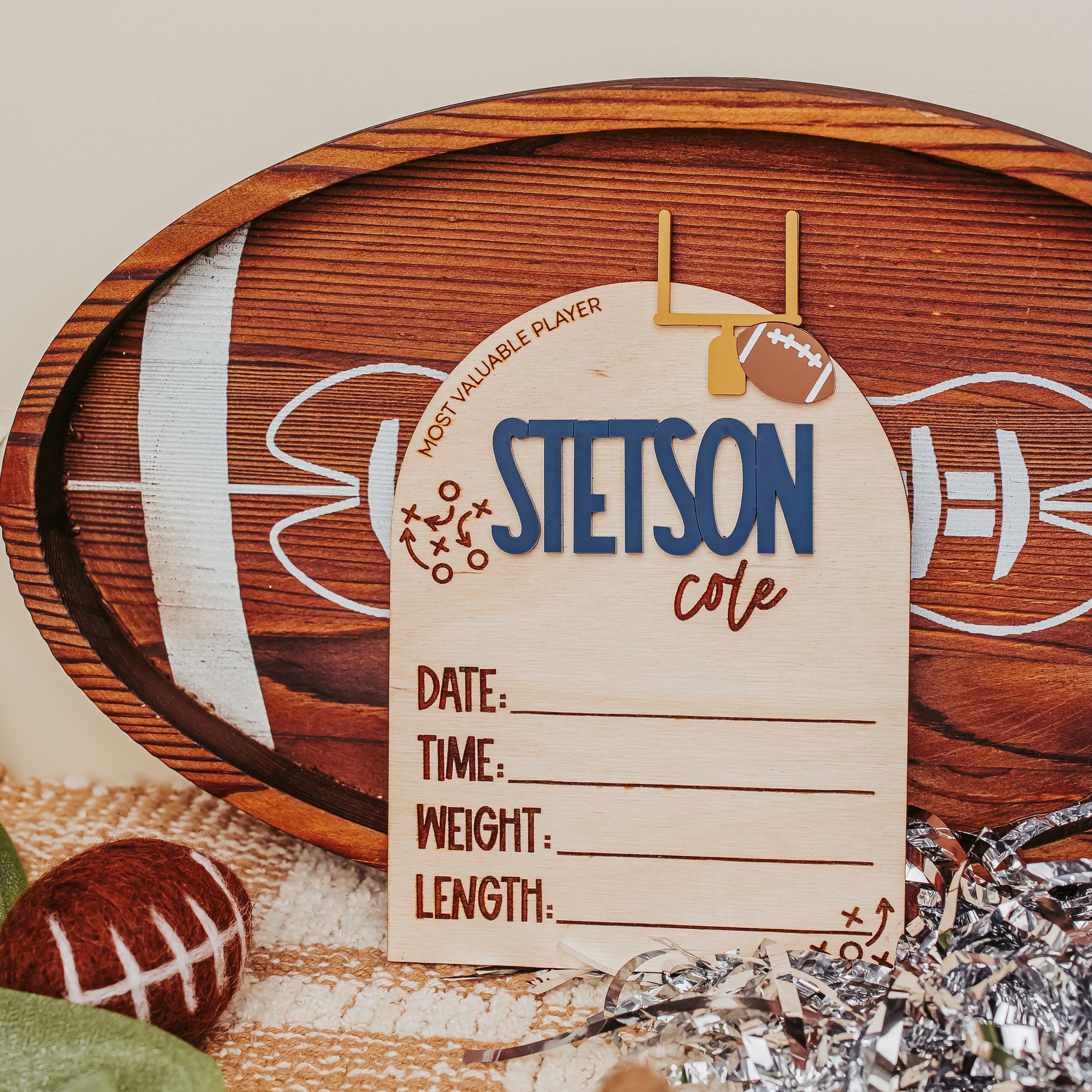 A wooden football-themed photo frame with a personalized name card reading 'STETSON cole' and sports fields printed on the wood, styled with a football, green fabric, and silver tinsel on a neutral background.