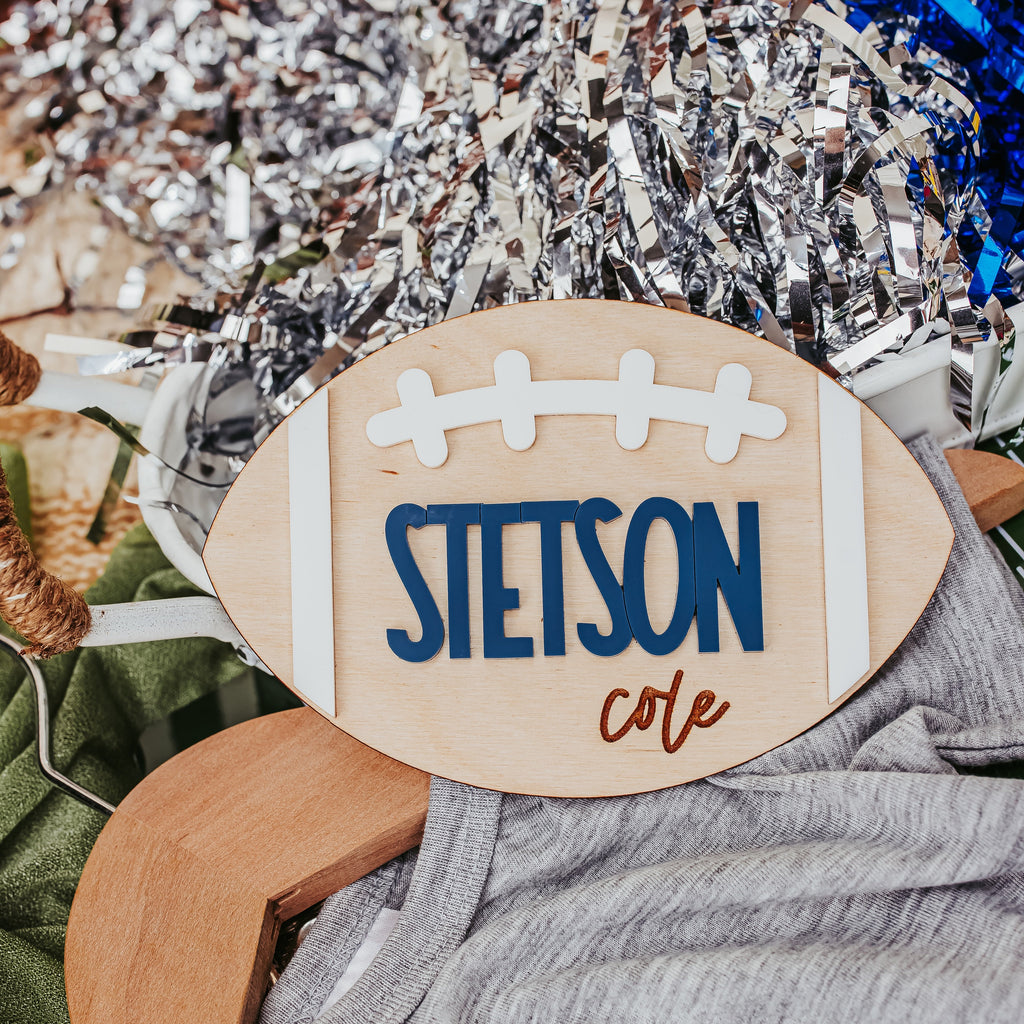 A custom wooden football-shaped baby announcement sign with the name 'STETSON cole' in blue and brown lettering, styled on a wooden hanger with a gray t-shirt and silver tinsel in the background.