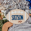 A custom wooden football-shaped baby announcement sign with the name 'STETSON cole' in blue and brown lettering, styled on a wooden hanger with a gray t-shirt and silver tinsel in the background.