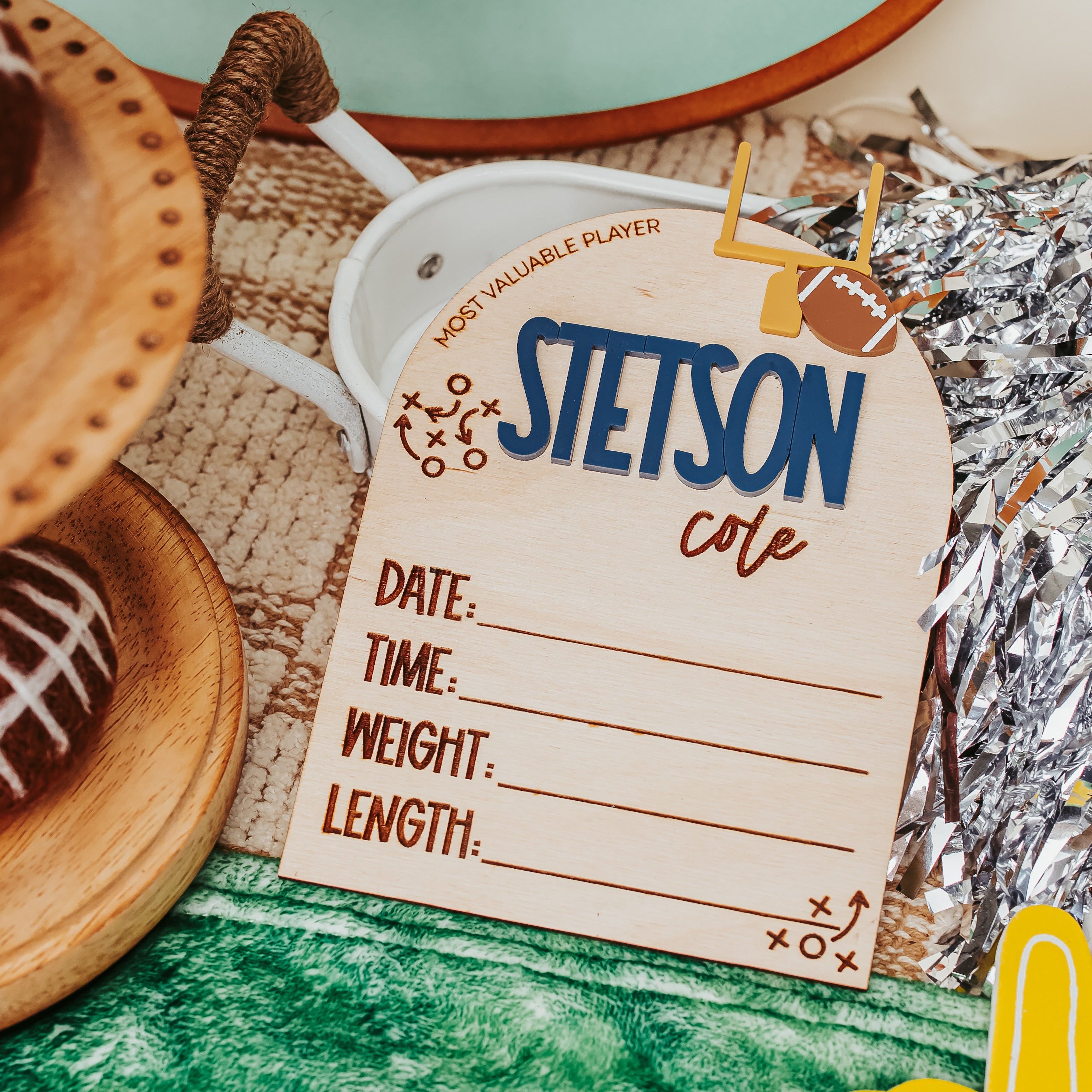 A football-themed party decoration set featuring a wooden name tag that reads 'STETSON cute' with lines for date, time, weight, and length, surrounded by a green turf mat, silver foil, a wooden football, and sports fan cutouts.