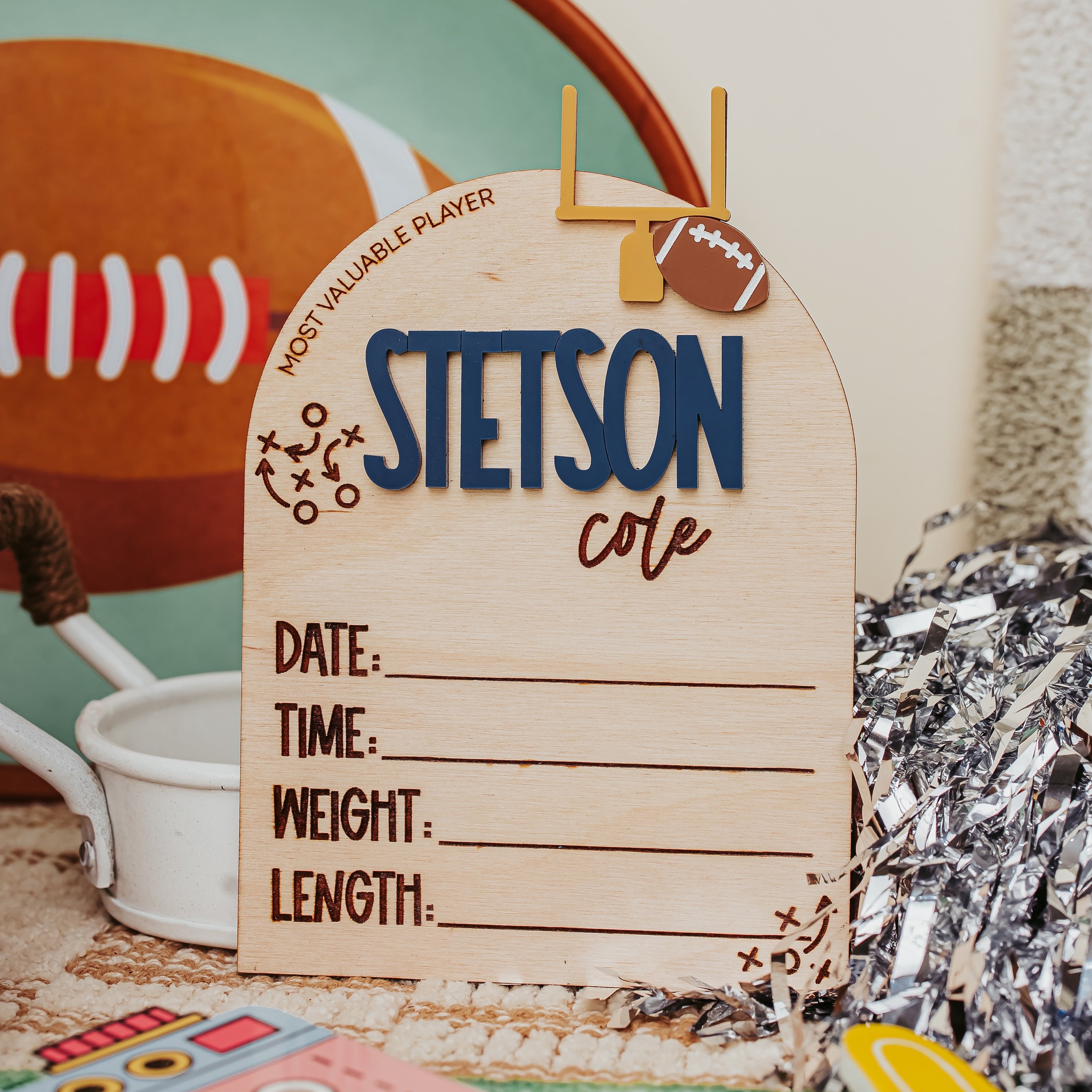 A personalized wooden baby stat sign for a football player, featuring the name 'STETSON cole' in blue and brown lettering, with fields for date, time, weight, and length. The sign is styled on a table with football-themed props including a football, a white watering can, and silver crinkle paper.