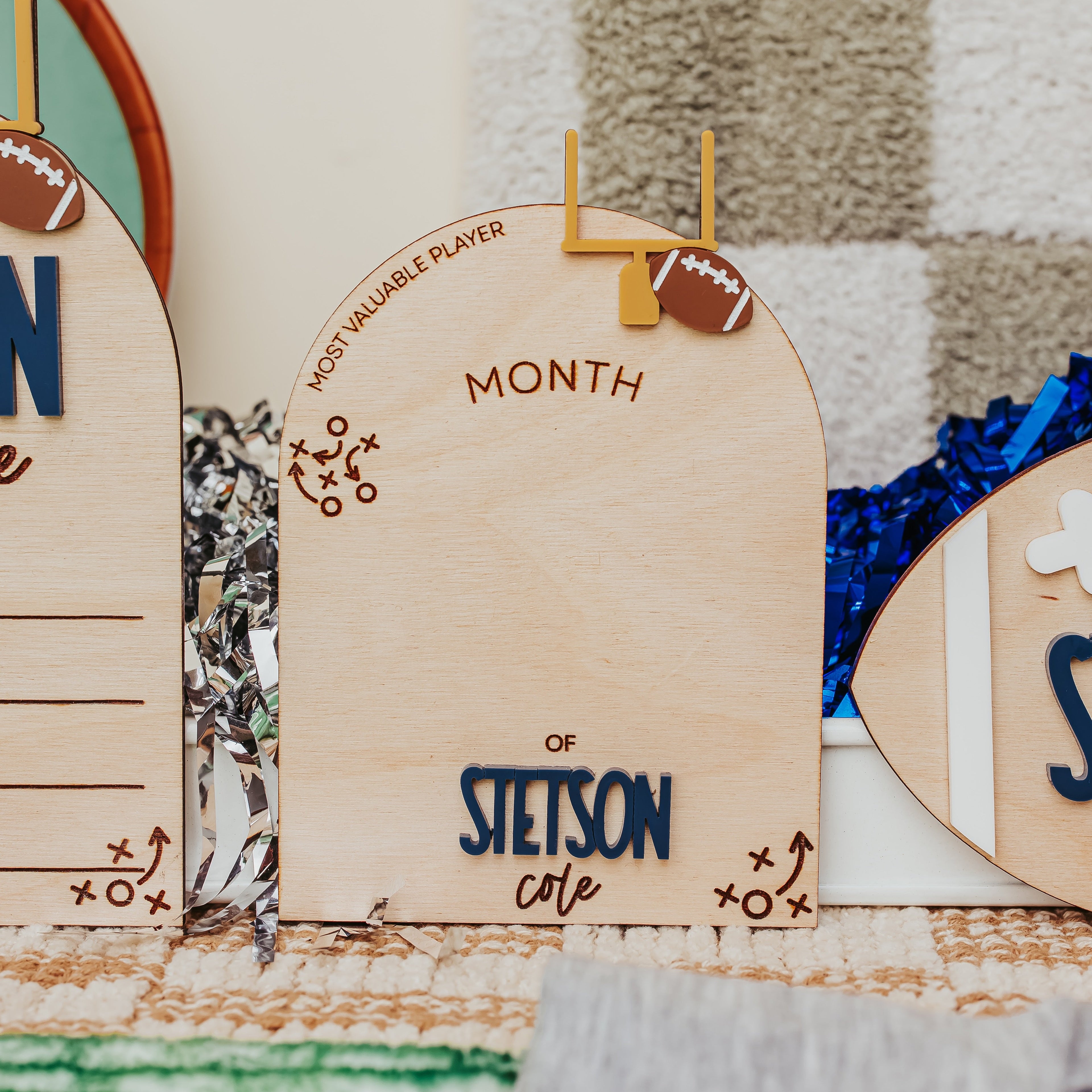 A close-up of a wooden arch baby milestone with football graphics and the text 'MOST VALUABLE PLAYER' and 'MONTH OF STETSON cote', shown on a decorated tabletop with other partial signs and festive ribbons.