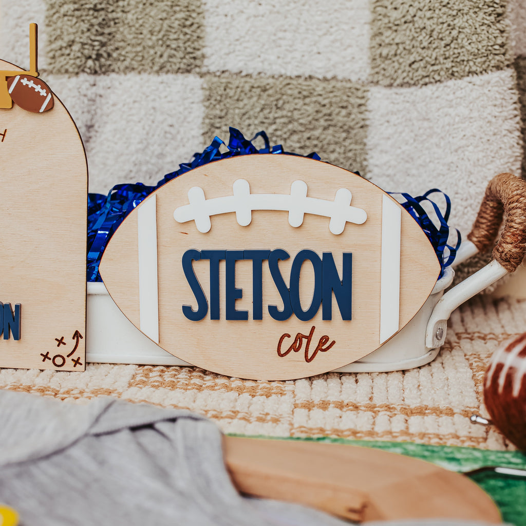 A wooden football-shaped sign with the name 'STETSON' in blue and 'cole' in brown script, styled on a checkered blanket with baby accessories.