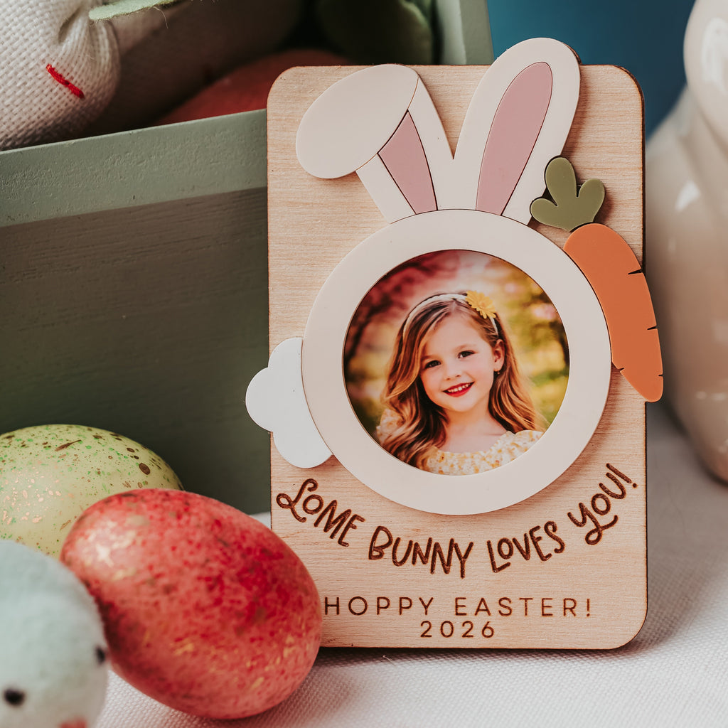 A personalized wooden Easter photo frame with bunny ears, a carrot accent, and a circular cutout showing a child's portrait. The frame is printed with the message 'LOVE BUNNY LOVES YOU! HOppy EASTER! 2026' and styled on a tabletop with decorative Easter eggs and bunny-themed items in the background.