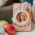 A personalized wooden Easter photo frame with bunny ears, a carrot accent, and a circular cutout showing a child's portrait. The frame is printed with the message 'LOVE BUNNY LOVES YOU! HOppy EASTER! 2026' and styled on a tabletop with decorative Easter eggs and bunny-themed items in the background.