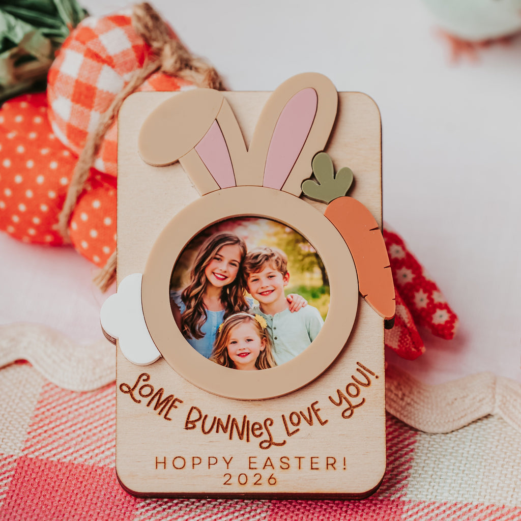 A personalized wooden Easter photo frame with bunny ears, a carrot accent, and a circular cutout showing a family photo. The frame is engraved with 'Some Bunnies Love You' and 'Hoppy Easter 2026', resting on a red and white gingham tablecloth with decorative Easter items in the background.