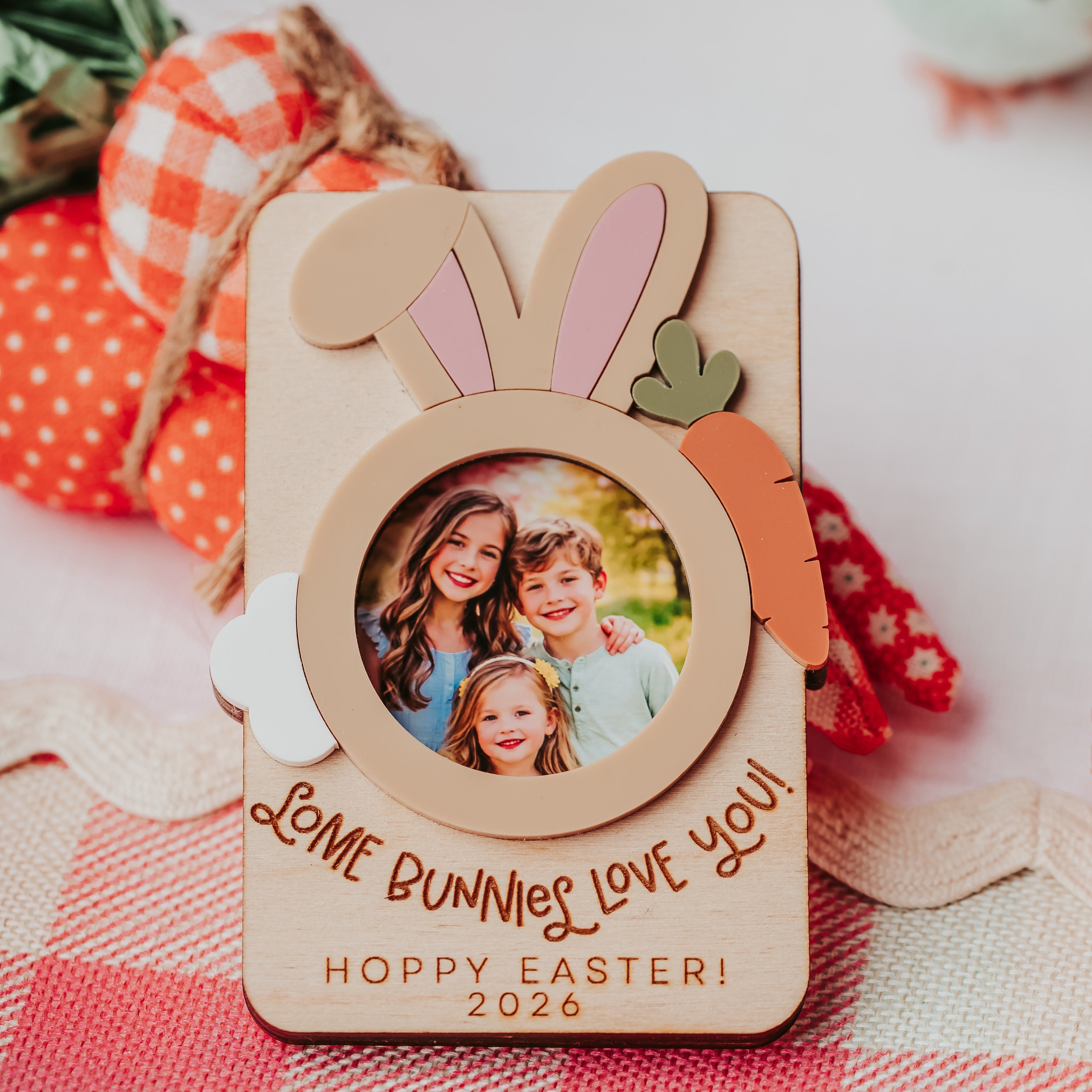 A personalized wooden Easter photo frame with bunny ears, a carrot accent, and a circular cutout showing a family photo. The frame is engraved with 'Some Bunnies Love You' and 'Hoppy Easter 2026', resting on a red and white gingham tablecloth with decorative Easter items in the background.