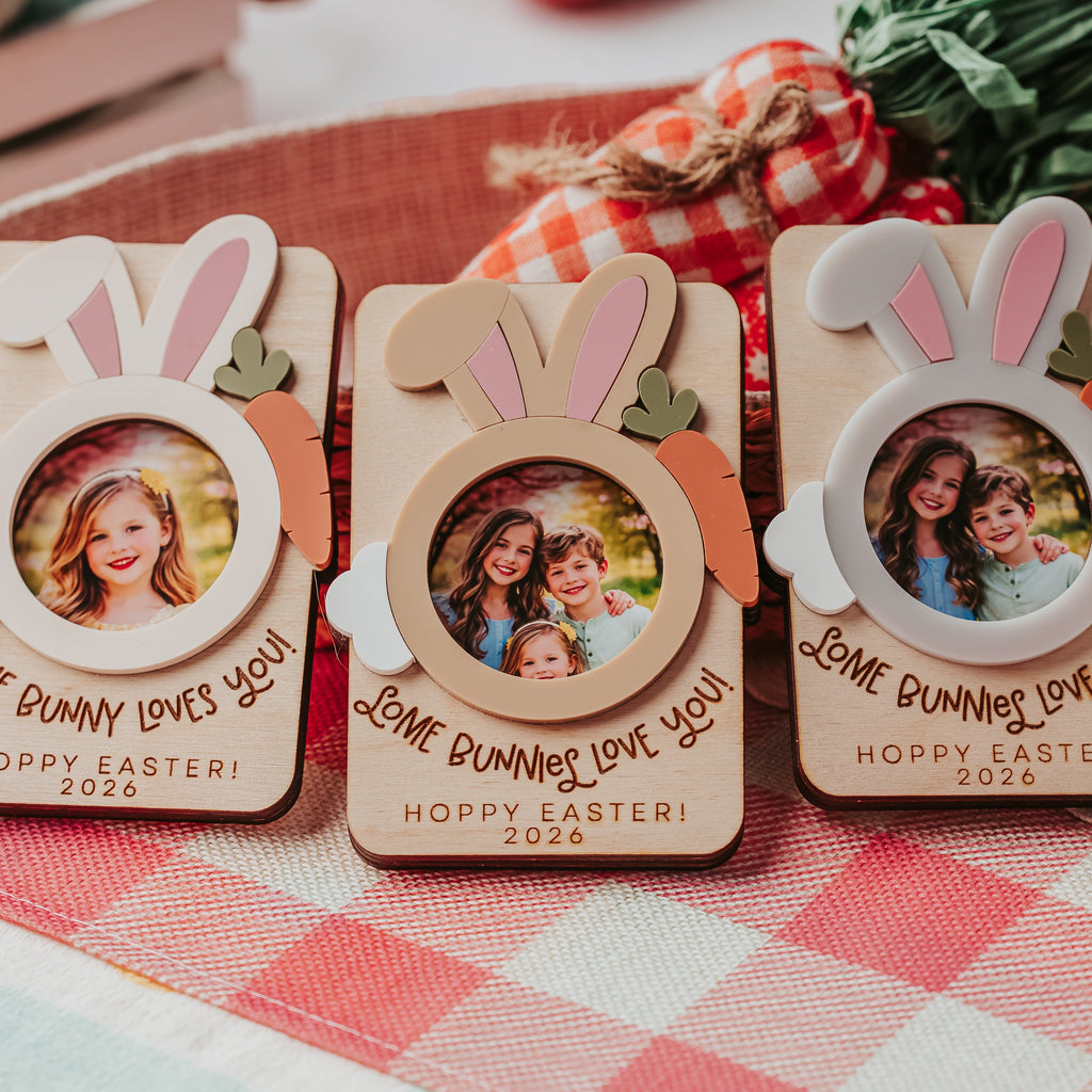 Three personalized wooden Easter photo frames with bunny ears and carrot accents, displayed on a red-and-white checkered tablecloth with Easter decorations in the background.