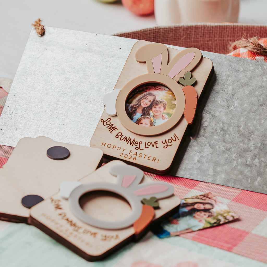 Easter-themed wooden photo frames with bunny ears and carrot accents, displayed on a table with a textured metal tray, a ceramic bunny figurine, and fruit in the background.