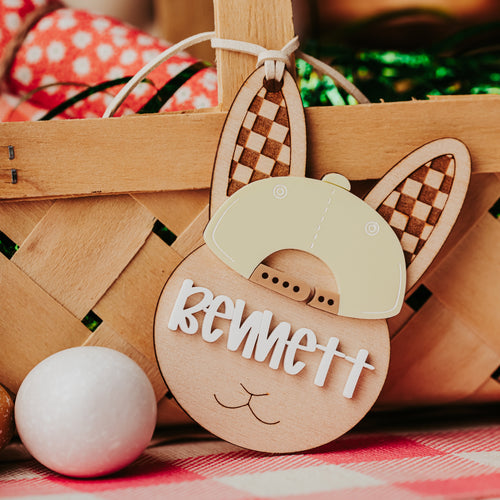 A personalized wooden bunny face easter basket tag with the name 'Bennett' in white script, featuring a beige bunny hat and checkered ears, resting in front of a woven basket on a red and white checkered blanket.