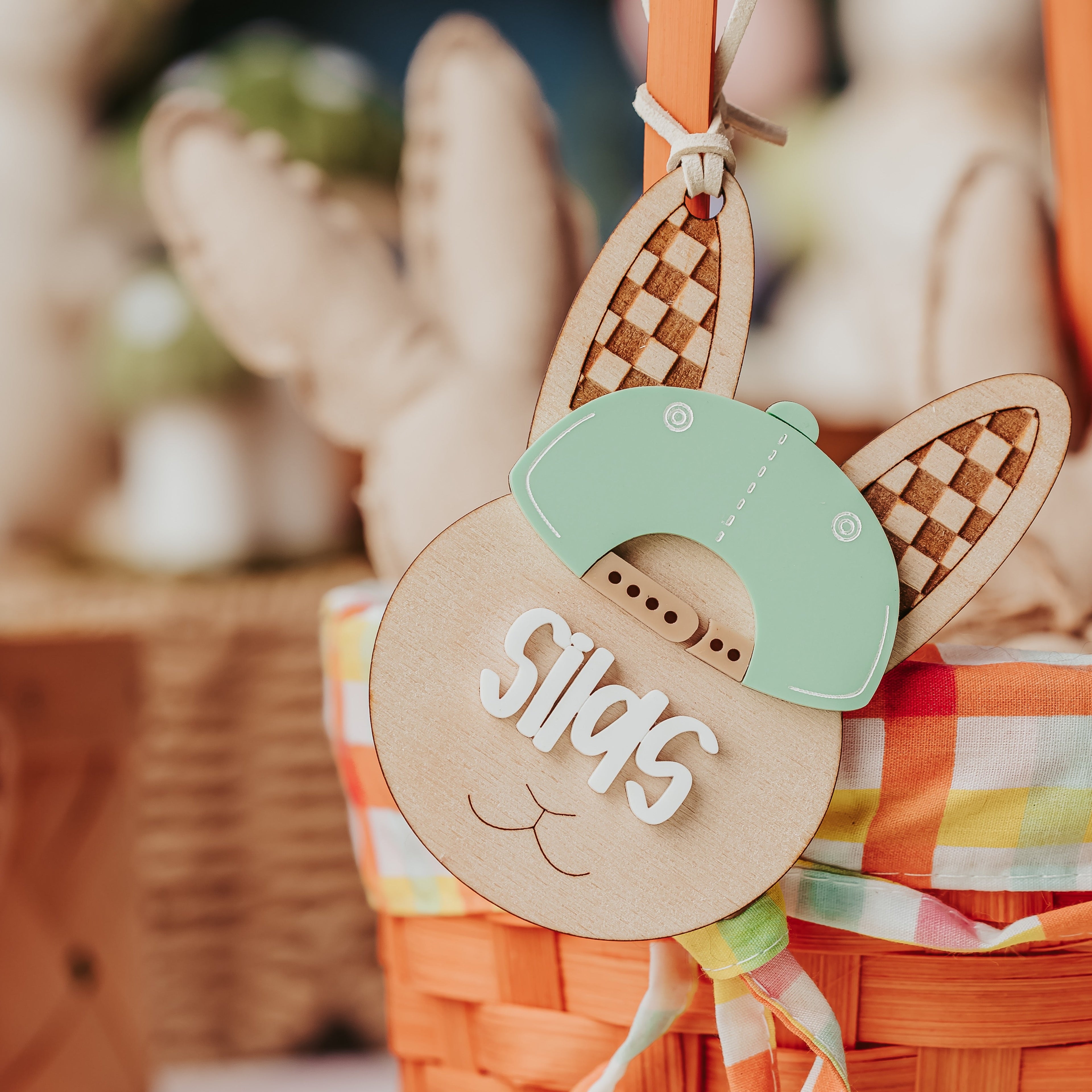 A close-up of a woven orange Easter basket with a wooden bunny-shaped easter basket tag that says 'Silas' in white script, decorated with a mint green hat and plaid ribbon, with bunny figures in the background.