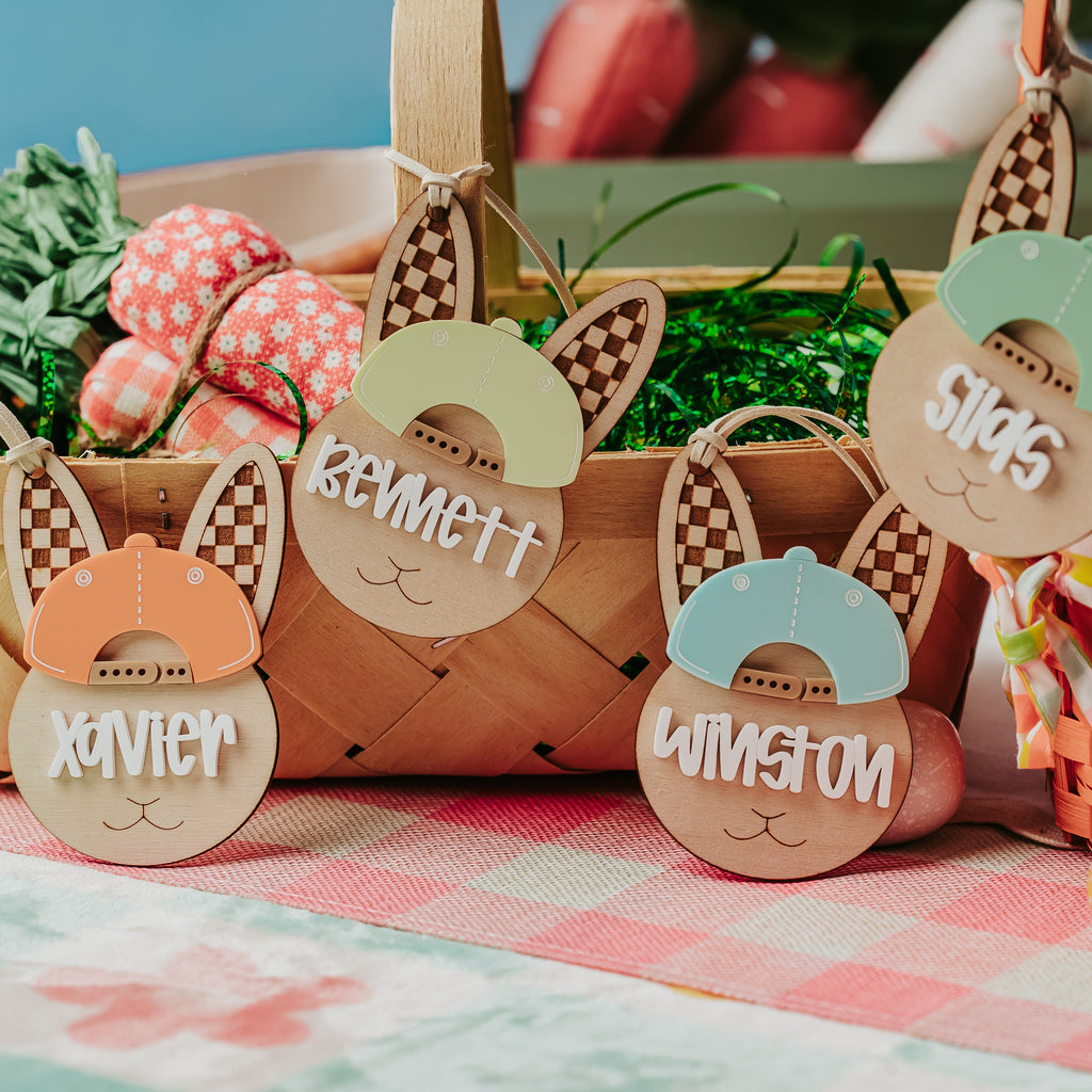 A styled Easter table scene with woven baskets, pastel eggs, green grass filler, and personalized bunny-shaped easter basket tags reading Xavier, Bennett, St. Louis, and Winston on a pink-and-white checkered cloth.