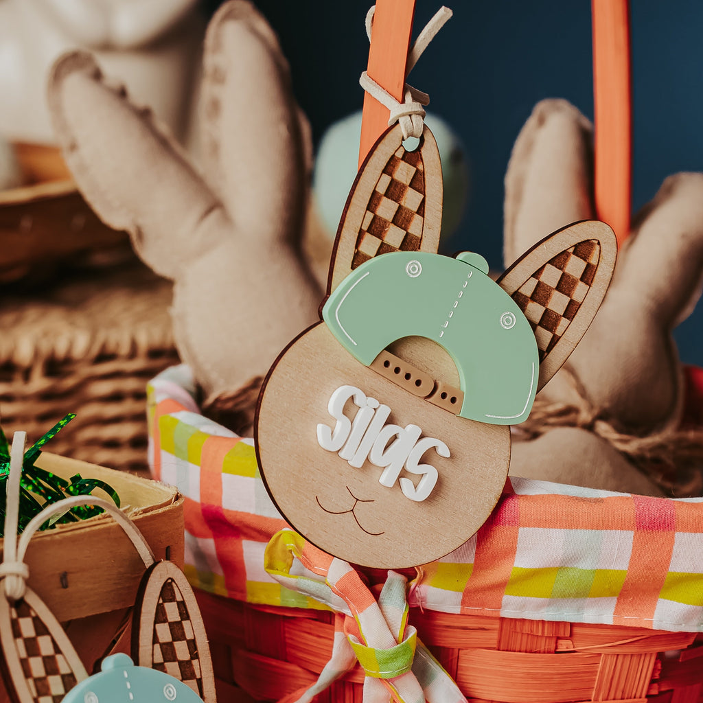 Easter-themed wooden bunny egg tags with cutout ears, a mint green hat, and white name cutouts, displayed in a woven basket with a red-and-white checkered lining and fabric.