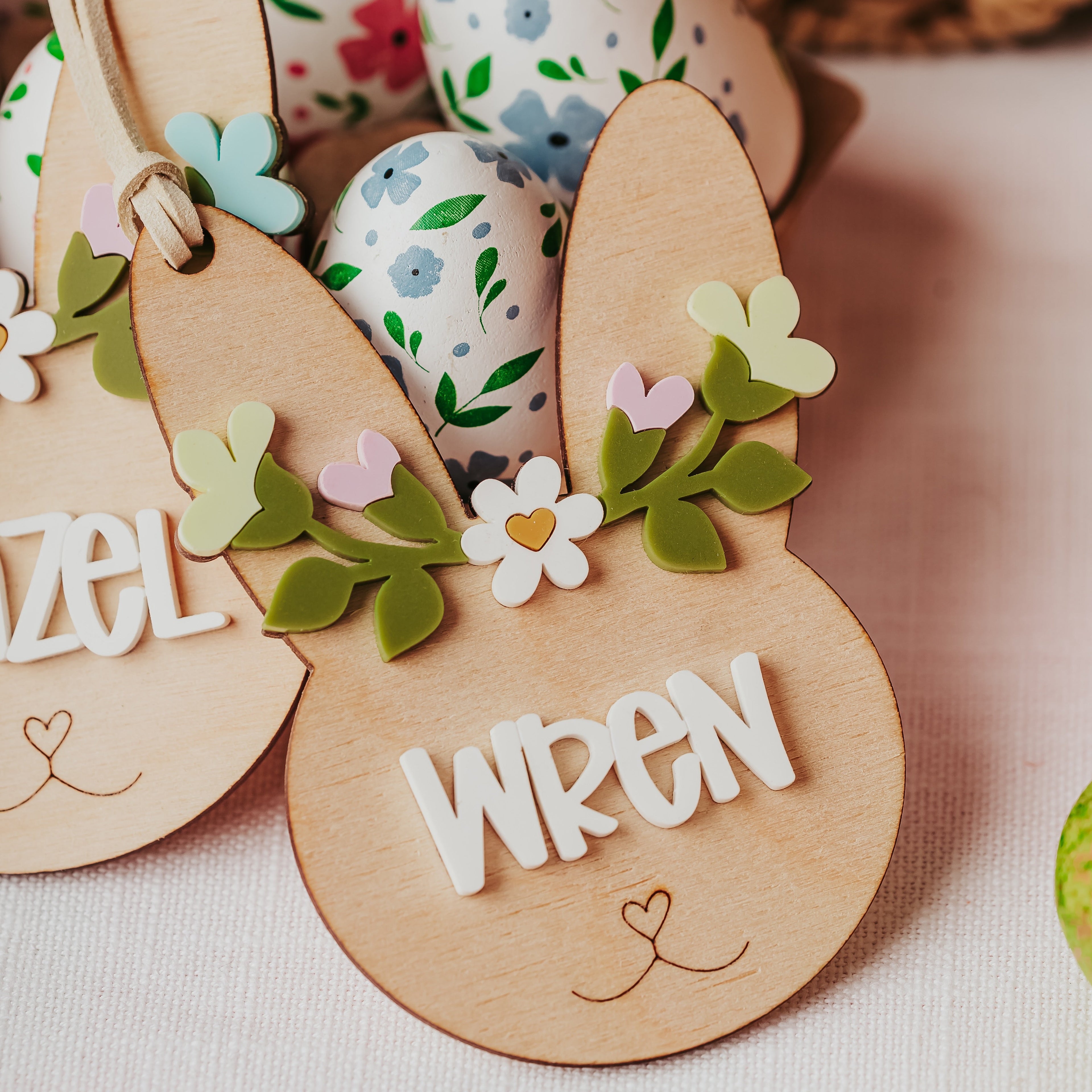 Personalized wooden bunny-shaped easter basket tag with floral accents and the name 'WREN' in white lettering, displayed among Easter eggs on a light fabric surface.
