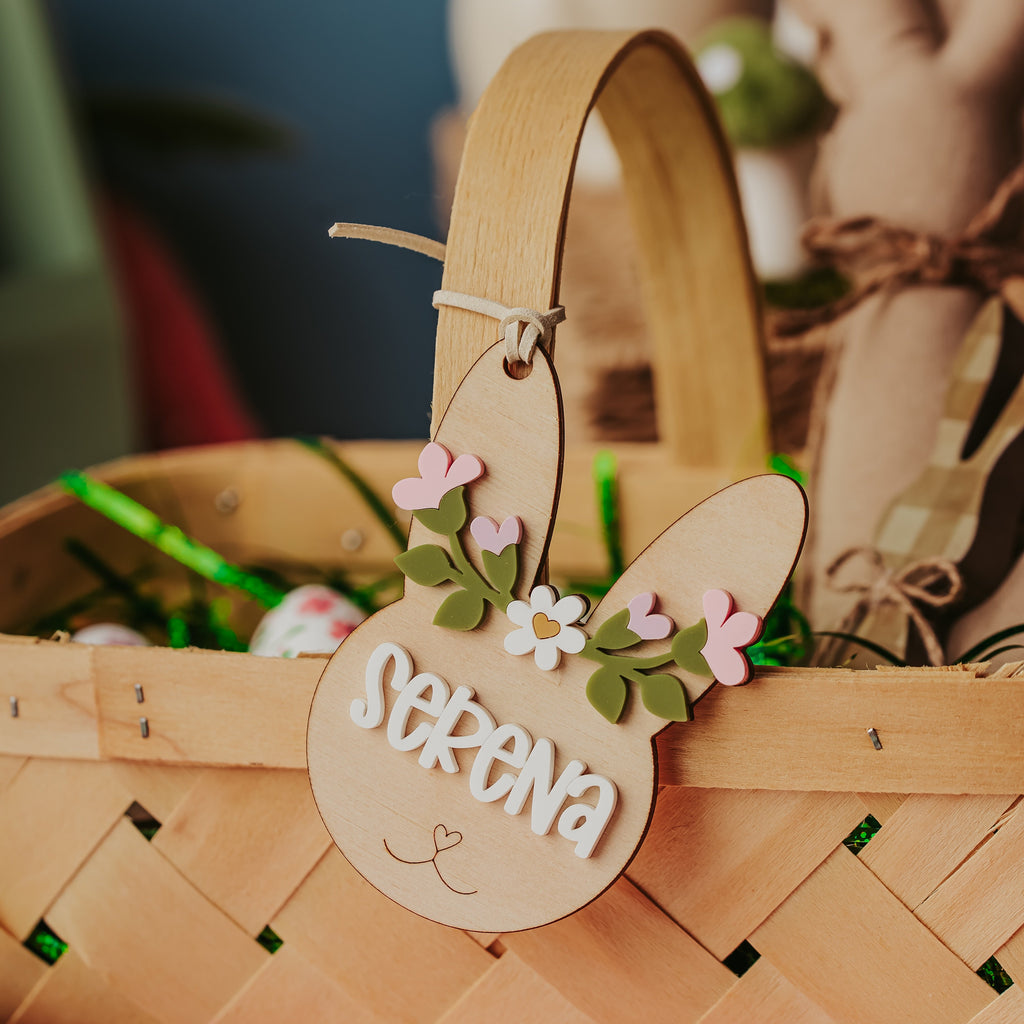 A woven natural wood Easter basket with a curved handle, decorated with a wooden bunny-shaped easter basket tag that reads 'SERENA' and has small pink and white flowers with green leaves. The basket is filled with green and white eggs and is set on a pink-and-white checkered tablecloth with a blurred rustic background.