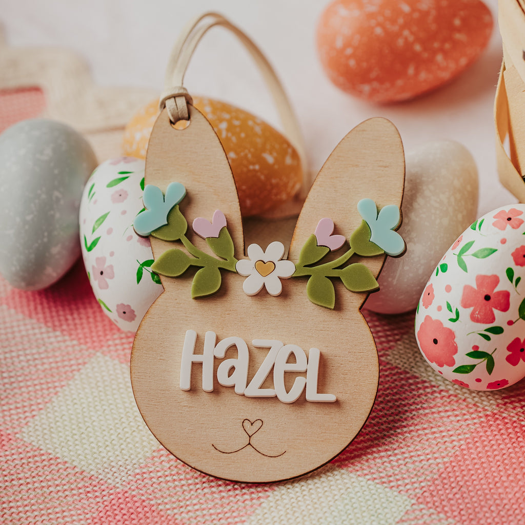 A personalized wooden bunny basket tag engraved with the name 'Hazel' and decorated with a small floral headband, styled among pastel Easter eggs on a pink-and-white checkered cloth.