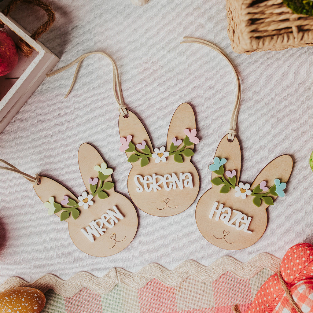Three personalized wooden bunny easter basket tags lay on a light fabric surface, decorated with small pastel flowers and the names 'Wren', 'Serena', and 'Hazel'. Colorful Easter eggs, a basket, and spring-themed props surround the ornaments.