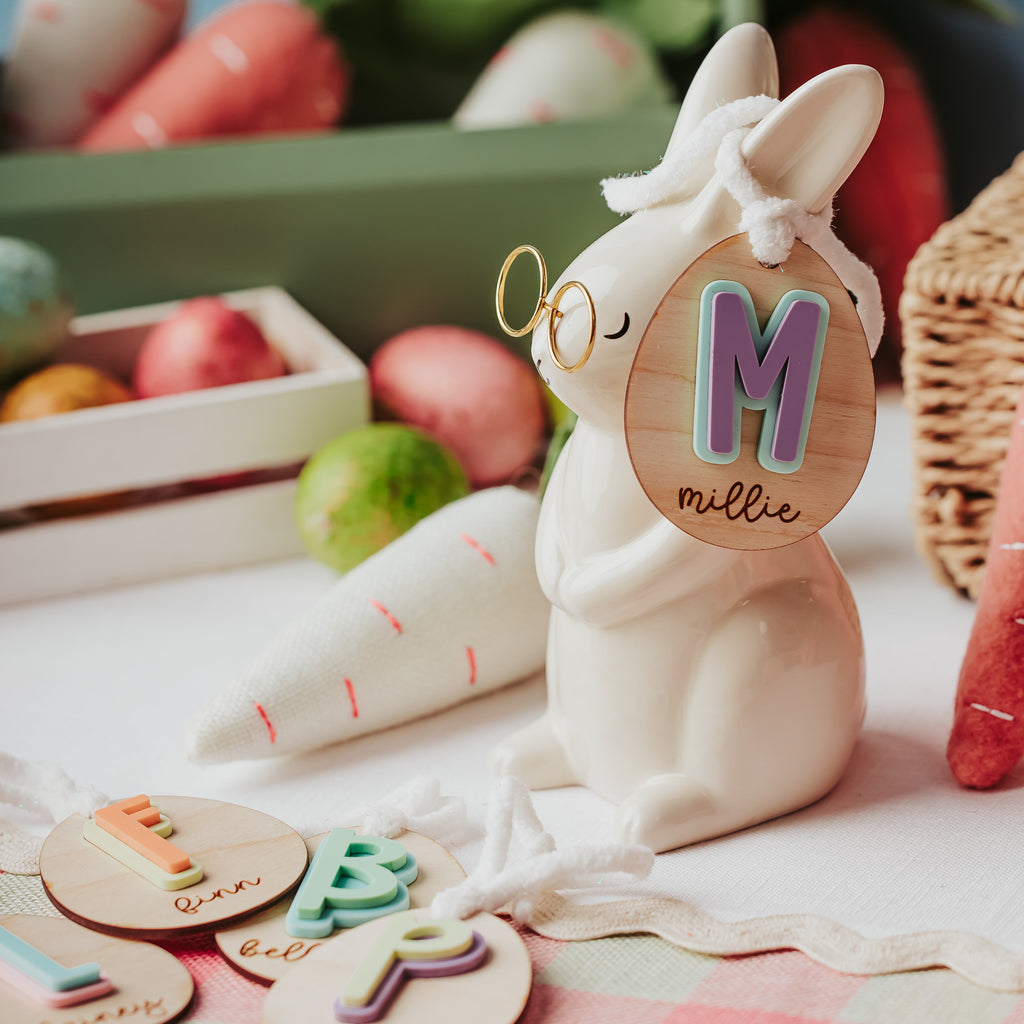 A ceramic Easter bunny figurine wearing gold-rimmed glasses and a white bow, holding a round wooden easter basket tag with a colorful letter 'M' and the name 'mille'. The bunny is styled on a pink-and-white checkered tablecloth with Easter-themed props including wooden letter cookies, a cone-shaped carrot, and a basket.