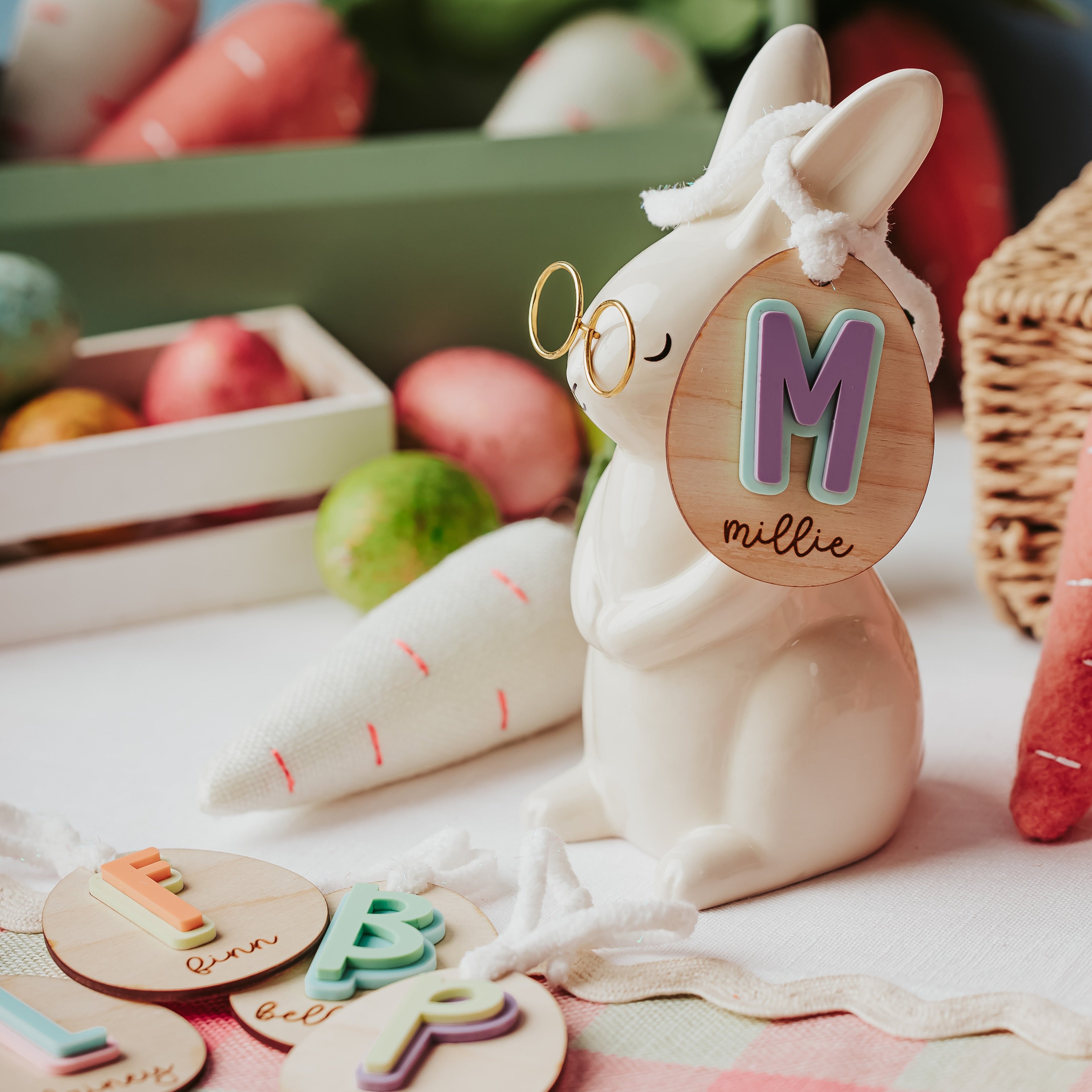 A ceramic Easter bunny figurine wearing gold-rimmed glasses and a white bow, holding a round wooden easter basket tag with a colorful letter 'M' and the name 'mille'. The bunny is styled on a pink-and-white checkered tablecloth with Easter-themed props including wooden letter cookies, a cone-shaped carrot, and a basket.