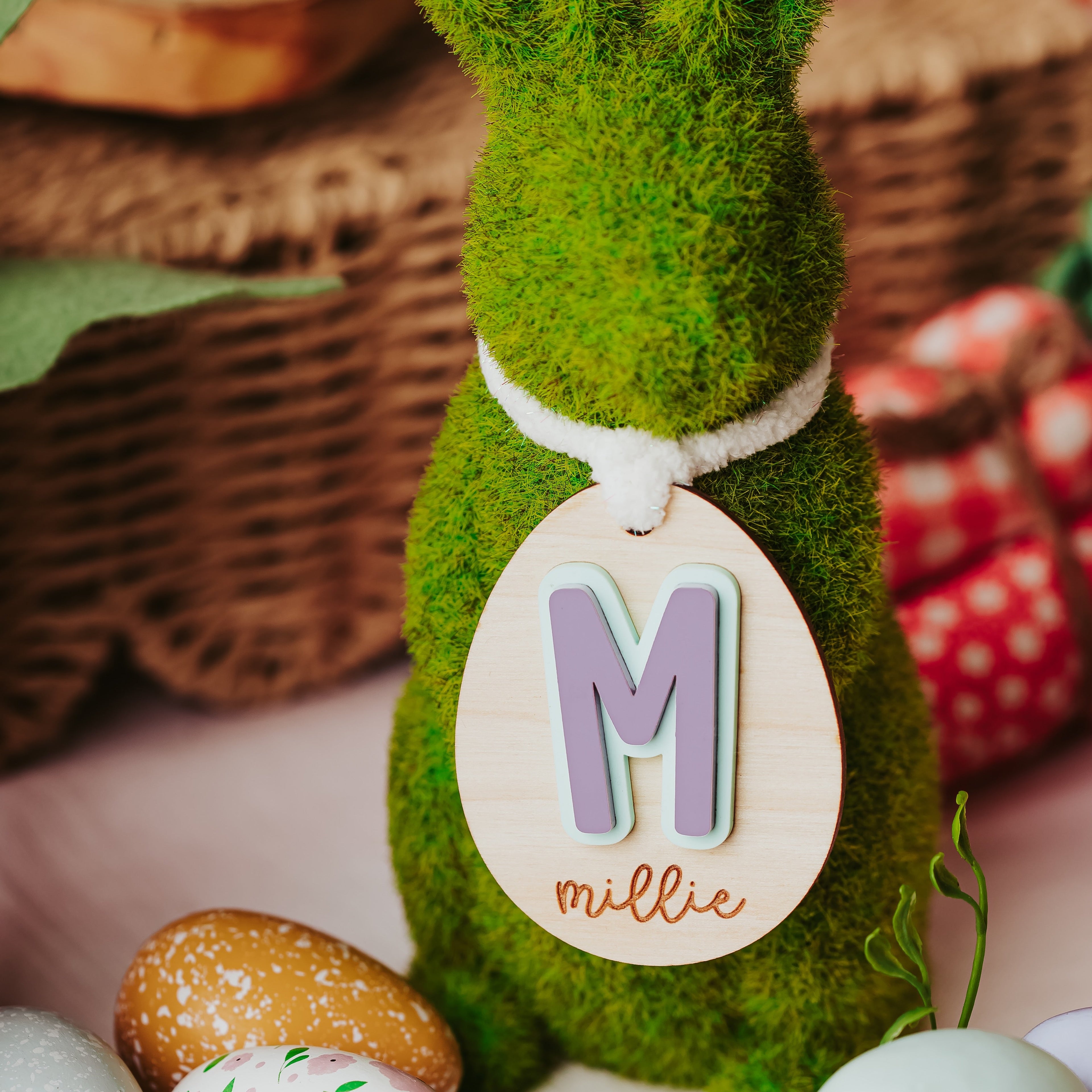 A green mossy bunny decoration with a white ribbon and a hanging wooden easter basket tag that reads 'M millie', styled with pastel Easter eggs and spring decor in a basket setting.