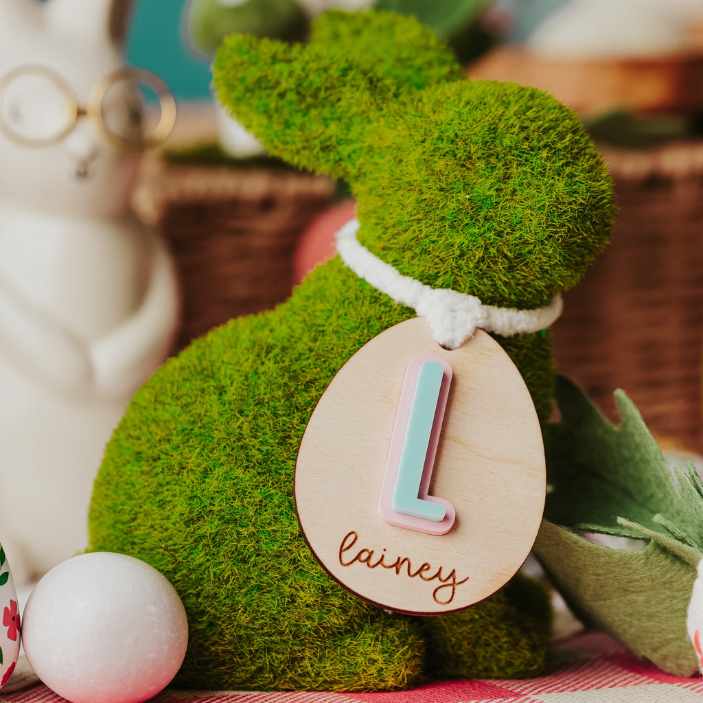 A green moss bunny figurine with a wooden personalized easter basket  tag reading 'L. lainey' on a pink and white checkered blanket, surrounded by Easter-themed props.