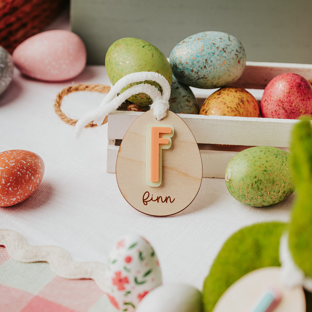 A styled Easter scene with colorful speckled and patterned eggs, a wooden crate, and two personalized wooden easter basket name tags hanging from white yarn. One tag shows a peach-colored letter 'F' and finn