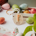 A styled Easter scene with colorful speckled and patterned eggs, a wooden crate, and two personalized wooden easter basket name tags hanging from white yarn. One tag shows a peach-colored letter 'F' and finn