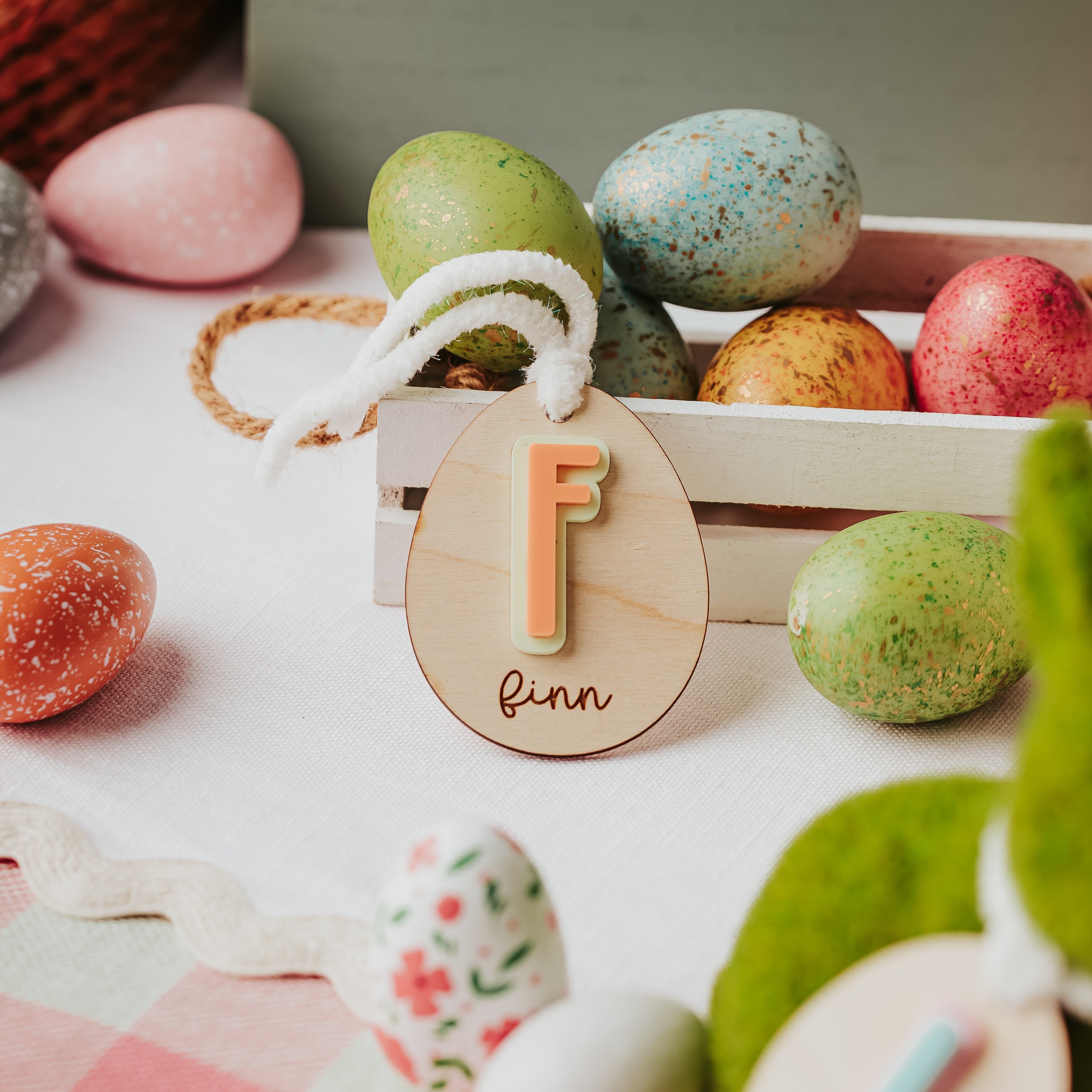 A styled Easter scene with colorful speckled and patterned eggs, a wooden crate, and two personalized wooden easter basket name tags hanging from white yarn. One tag shows a peach-colored letter 'F' and finn