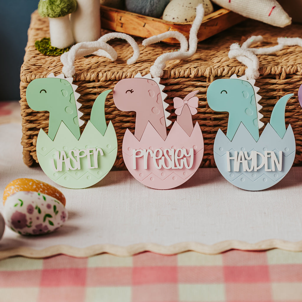Four personalized acrylic dinosaur egg basket tag in pastel green, pink, blue, and purple, displayed on a pink and white checkered tablecloth in front of a wicker basket.
