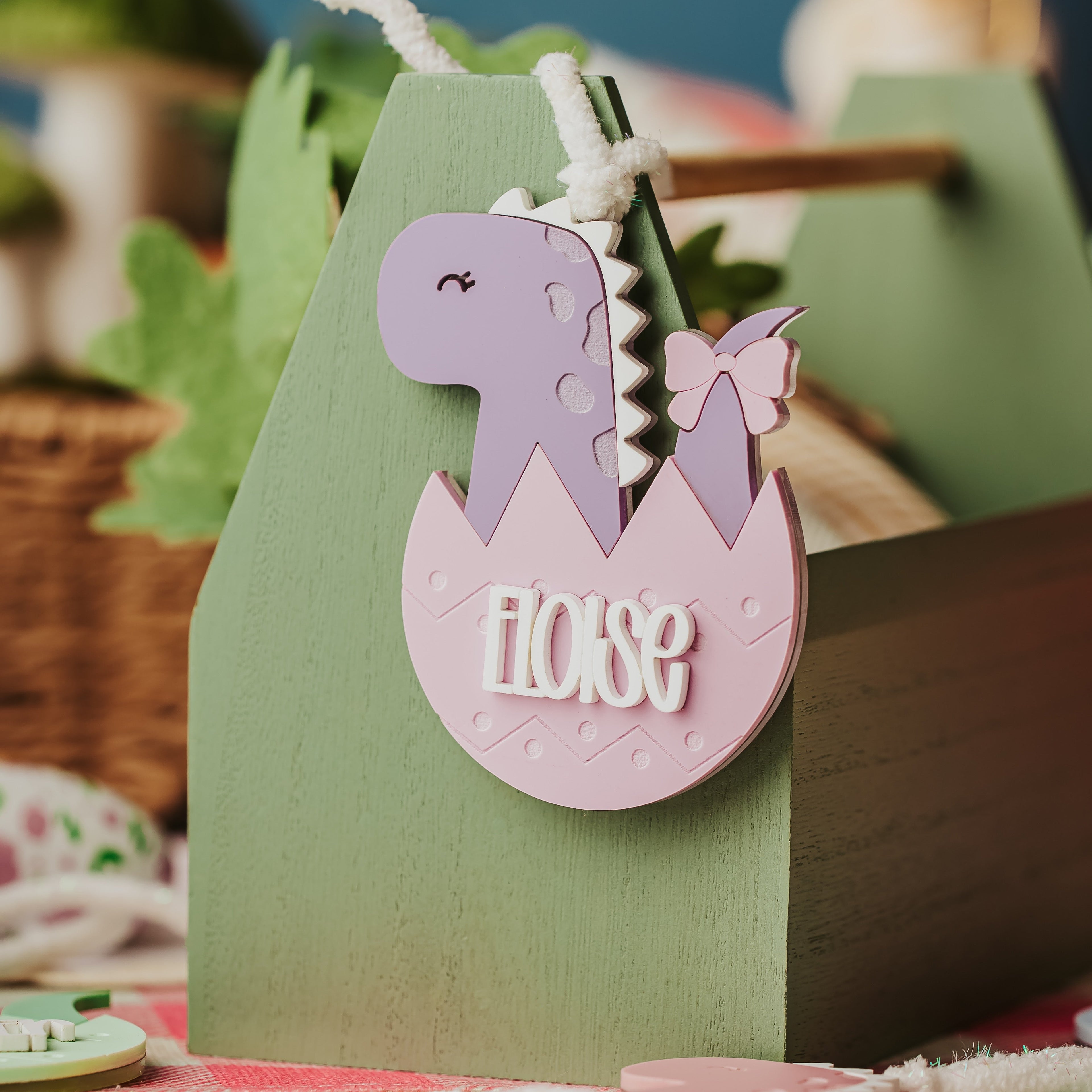 A close-up of a pastel green wooden Easter basket with a decorative hanging tag shaped like a purple dinosaur hatching from a pink egg, with the word 'ELOISE' on the tag.