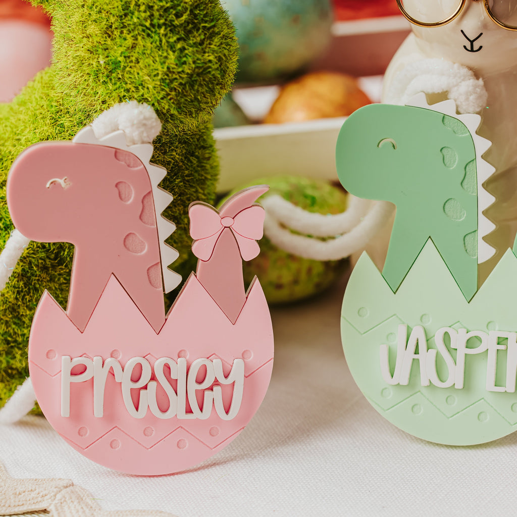 Two personalized dinosaur-shaped egg-shaped basket tags, one pink with the name 'Presley' and one mint green with the name 'Jasper', displayed on a styled Easter-themed surface with a bunny figurine and decorative eggs in the background.