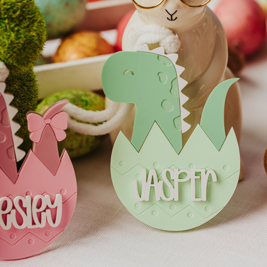 Personalized Easter egg cutouts on a table, featuring a green dinosaur hatching from an egg with the name 'JASPER' and part of a pink egg with the name 'PEESLEY'. A ceramic bunny figurine wearing round glasses stands behind them.