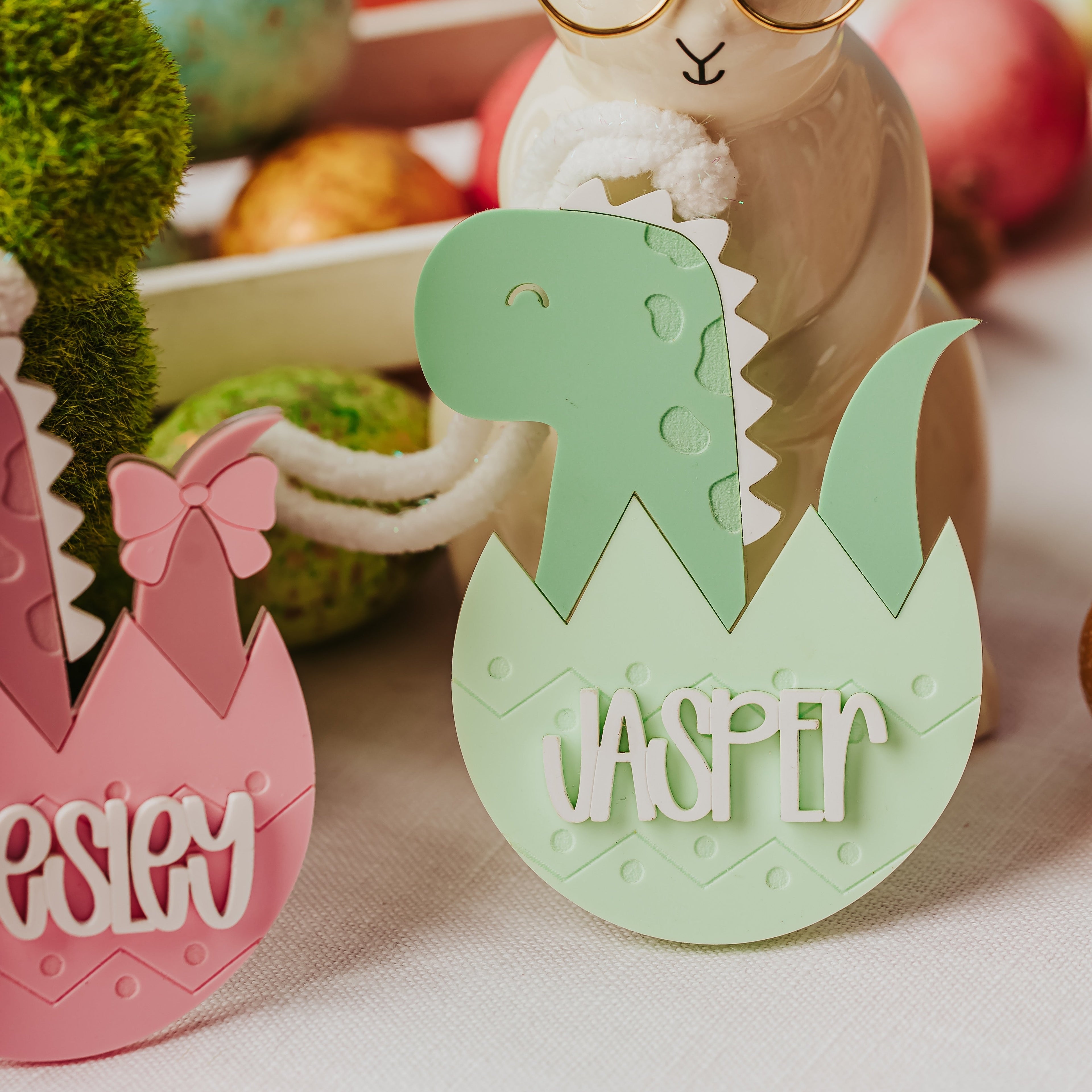 Personalized Easter egg cutouts on a table, featuring a green dinosaur hatching from an egg with the name 'JASPER' and part of a pink egg with the name 'PEESLEY'. A ceramic bunny figurine wearing round glasses stands behind them.