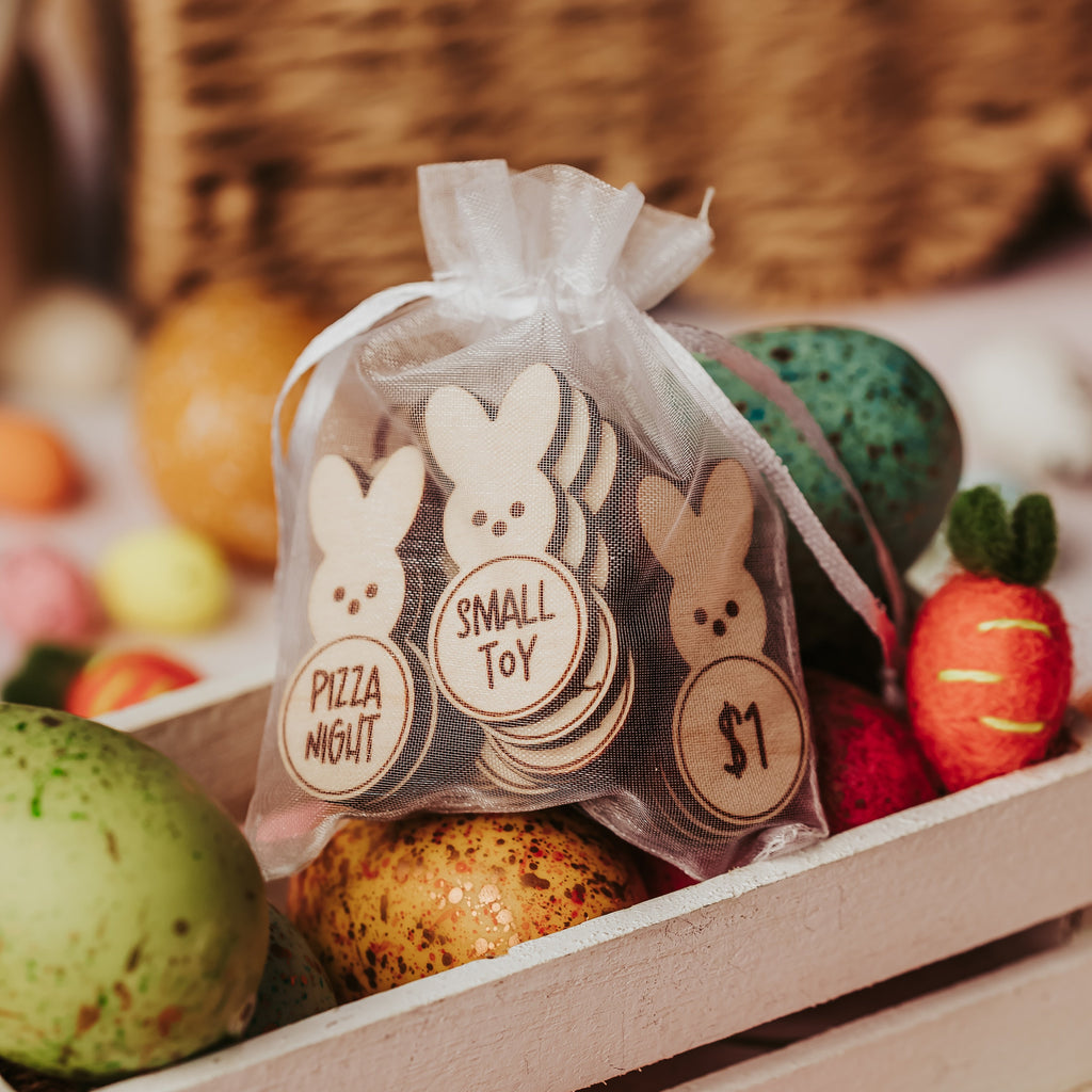 A small see-through white organza bag filled with easter egg bunny tokens  labeled 'PIZZA NIGHT', 'SMALL TOY', and '$1' sits in a white wooden crate with colorful eggs and felt carrots around it.