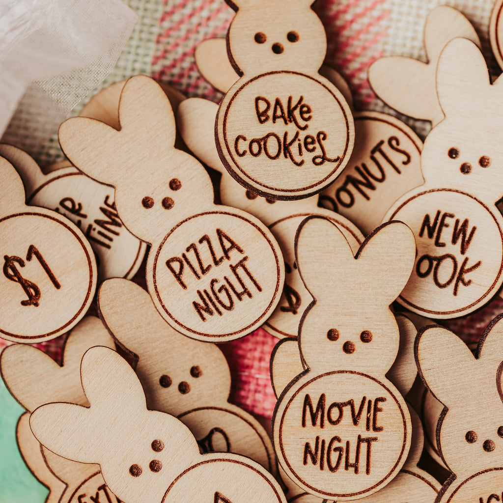 A pile of laser-cut wooden bunny-shaped easter egg tokens with various themed text options like 'Bake Cookies', 'Pizza Night', and 'Movie Night', displayed on a pink and white plaid blanket.