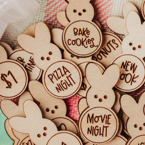 A pile of laser-cut wooden bunny-shaped easter egg tokens with various themed text options like 'Bake Cookies', 'Pizza Night', and 'Movie Night', displayed on a pink and white plaid blanket.