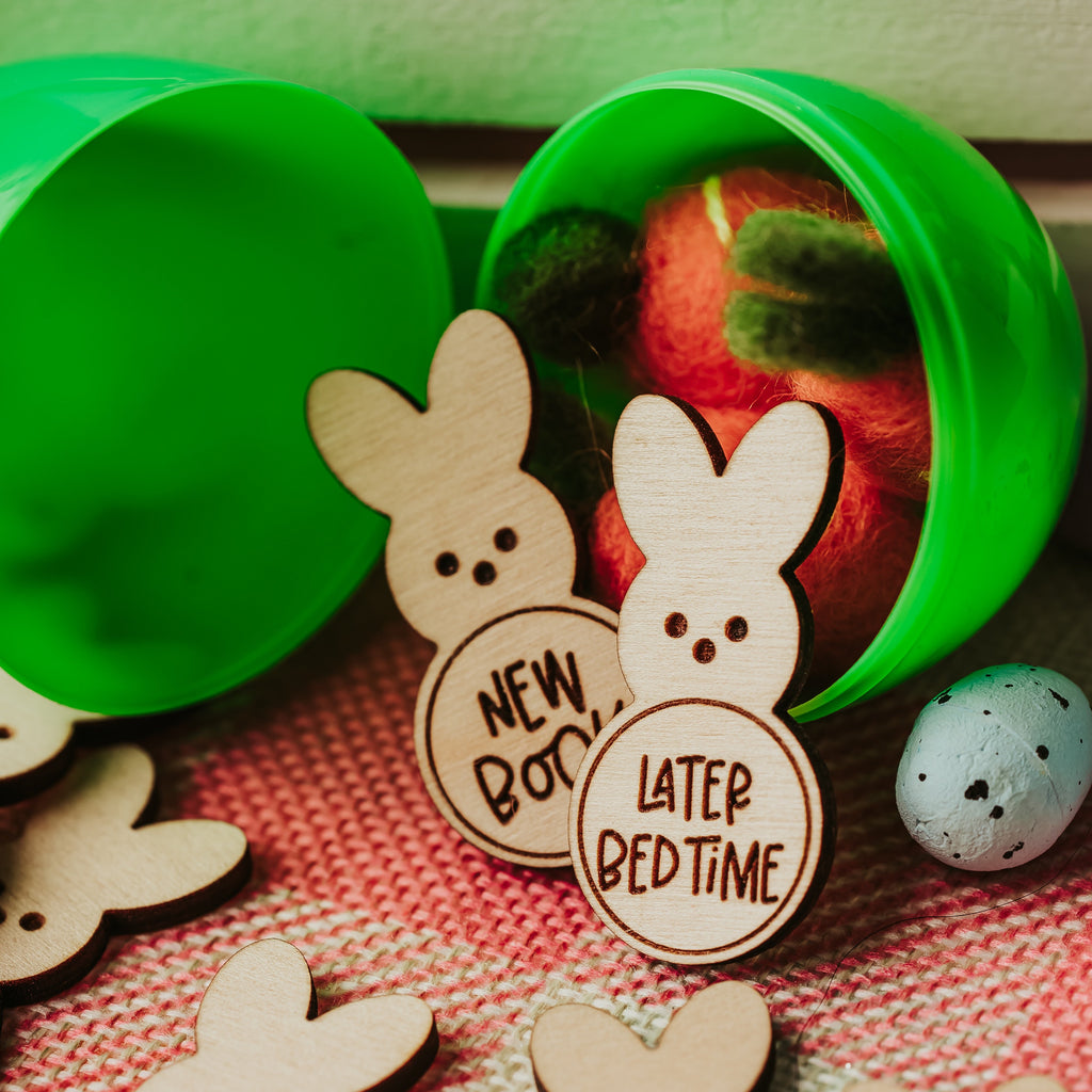 Easter-themed wooden bunny easter egg tokens  with engraved text like 'NEW BOOK', 'LATER BEDTIME', 'SMALL TOY', and '$1' arranged on a pink and white woven plaid surface, alongside a green plastic egg containing colorful items and a speckled egg.