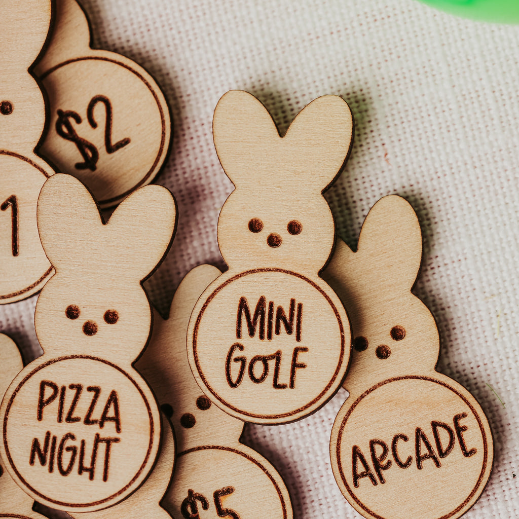 Close-up of small wooden bunny-shaped easter egg tokens with engraved round coins showing activities like pizza night, mini golf, arcade, and dollar amounts, arranged on a light fabric surface.