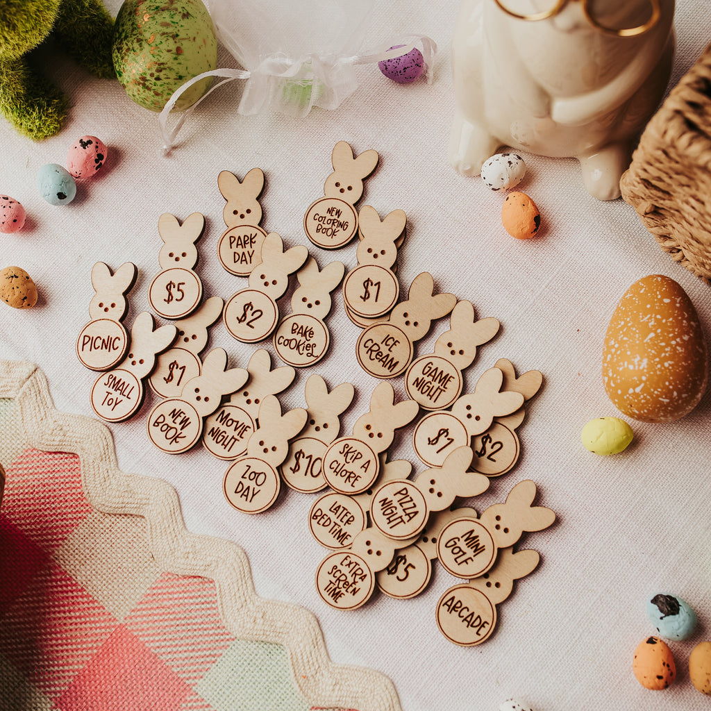 A styled flat-lay of small wooden bunny-shaped easter egg tokens engraved with Easter-themed phrases such as 'new book' or 'small toy' surrounded by pastel chocolate eggs, a cream bunny figurine, and a pink-and-white checkered fabric on a light surface.
