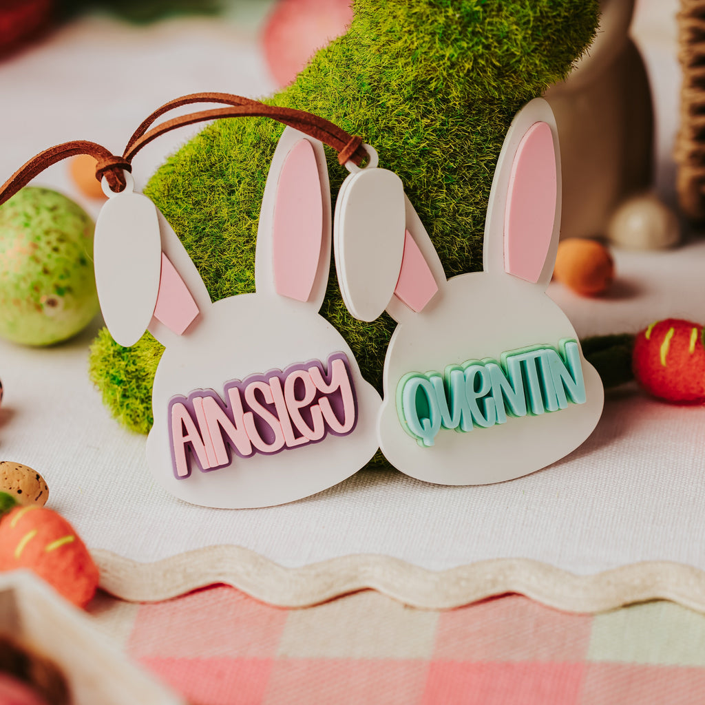 Two white bunny-shaped name basket tags with pink inner ears hang from a brown ribbon. The left plaque reads "ANISLEY" in pink letters and the right reads "QUENTIN" in teal letters.