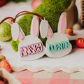 Two white bunny-shaped name basket tags with pink inner ears hang from a brown ribbon. The left plaque reads "ANISLEY" in pink letters and the right reads "QUENTIN" in teal letters.