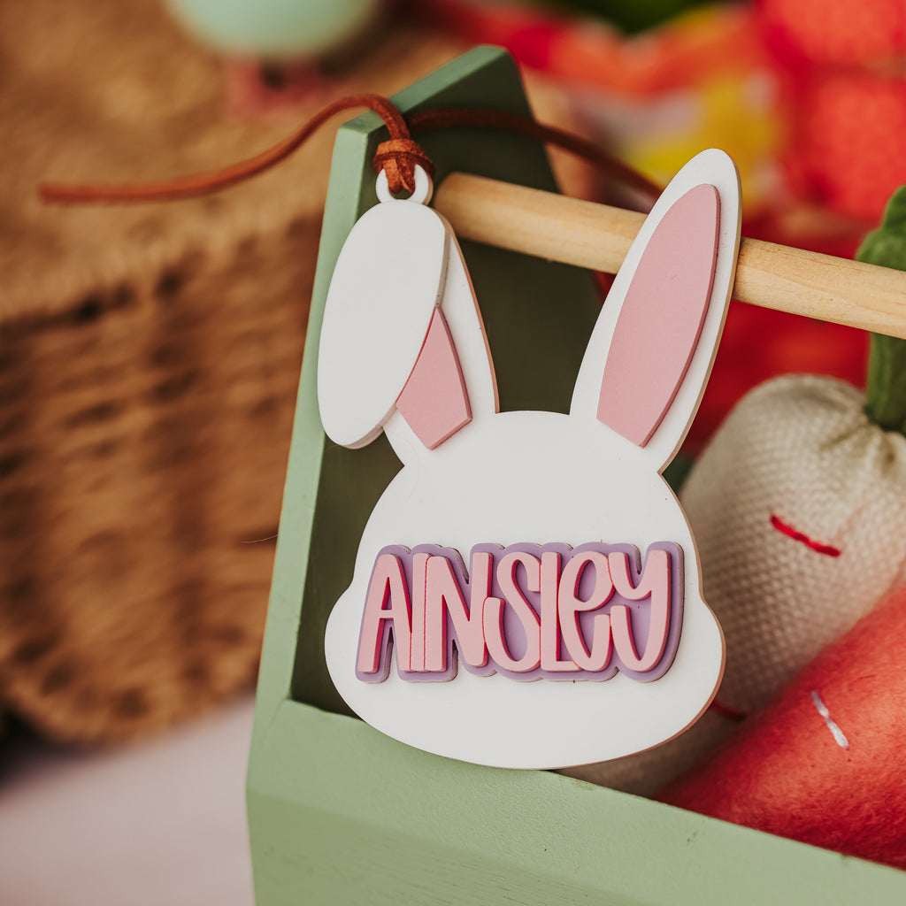 A personalized acrylic bunny-shaped basket tag with the name 'AINSLEY' in pink raised letters, hanging from a string in a decorative basket.