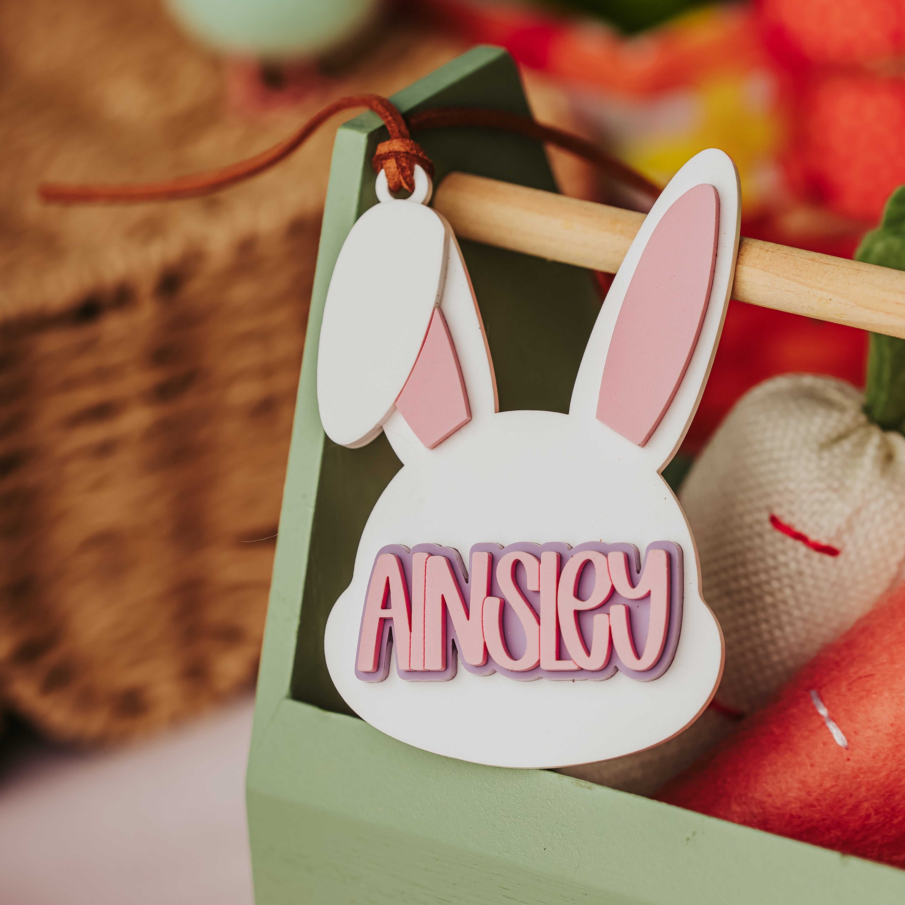 A personalized acrylic bunny-shaped basket tag with the name 'AINSLEY' in pink raised letters, hanging from a string in a decorative basket.