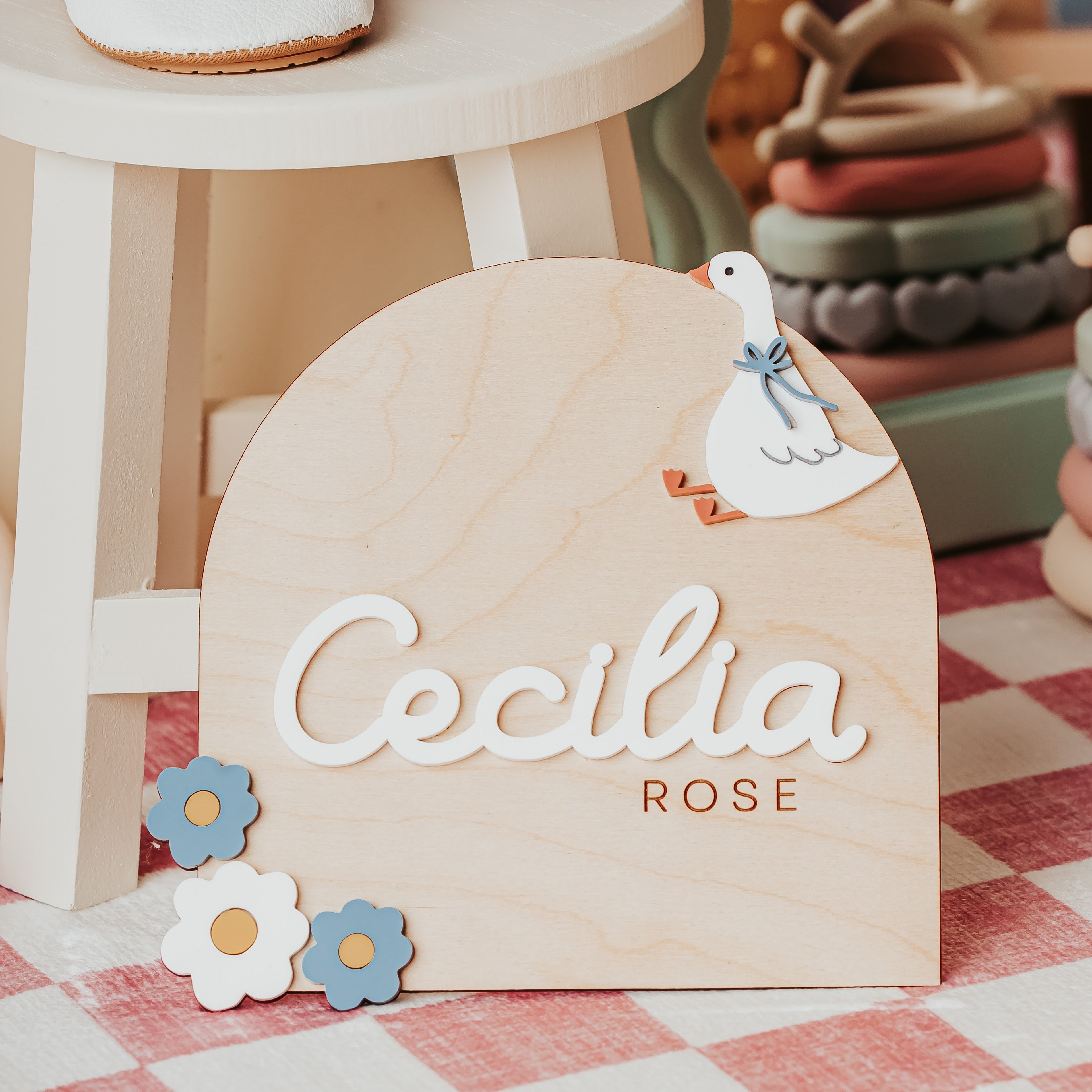 A personalized silly goose wooden baby sign with the name 'Cecilia' in raised white script and 'ROSE' below in gold, decorated with a white dove and small flower cutouts, displayed on a checkered floor next to a white stool.