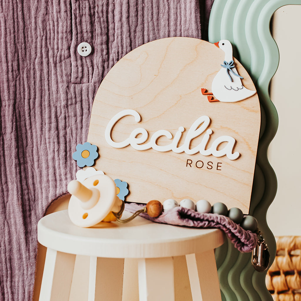 A silly goose wooden baby name announcement keepsake sign with the name 'Cecilia ROSE' in white script, styled on a small white stool with baby accessories against a mauve ribbed fabric backdrop.