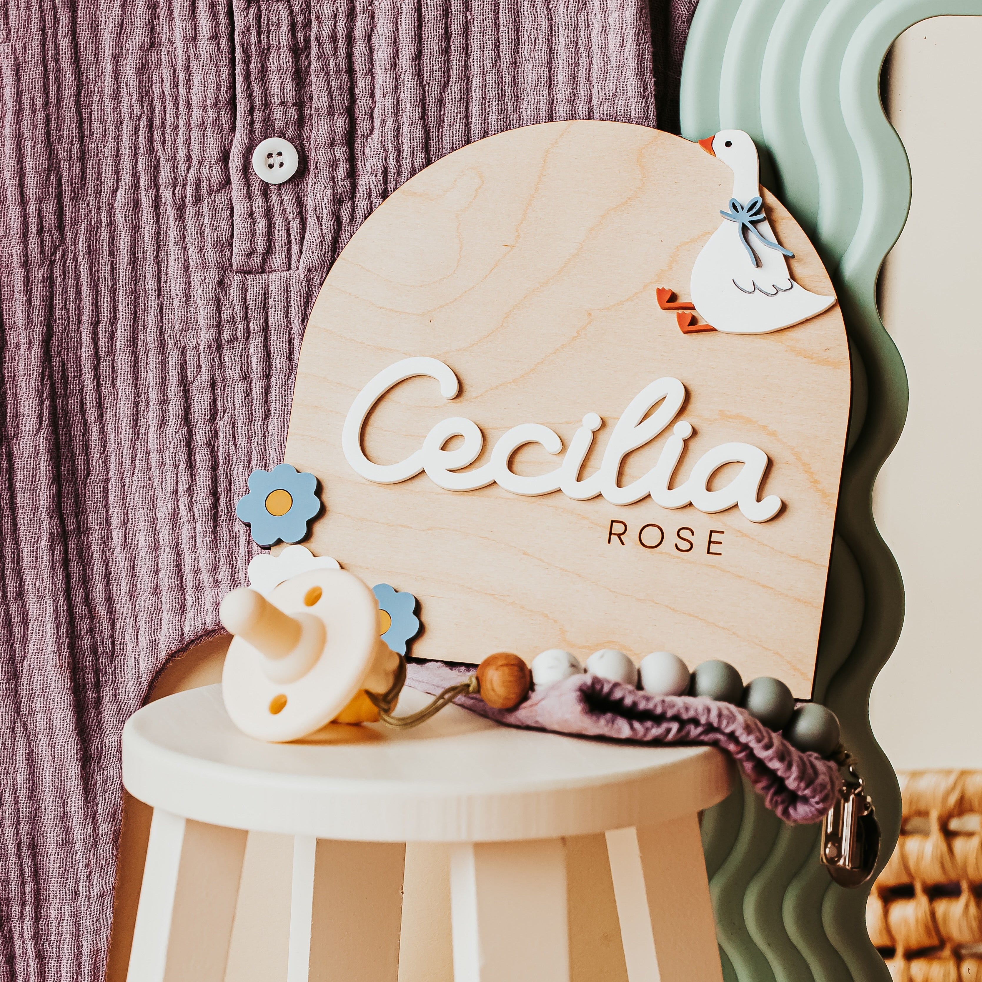 A silly goose wooden baby name announcement keepsake sign with the name 'Cecilia ROSE' in white script, styled on a small white stool with baby accessories against a mauve ribbed fabric backdrop.