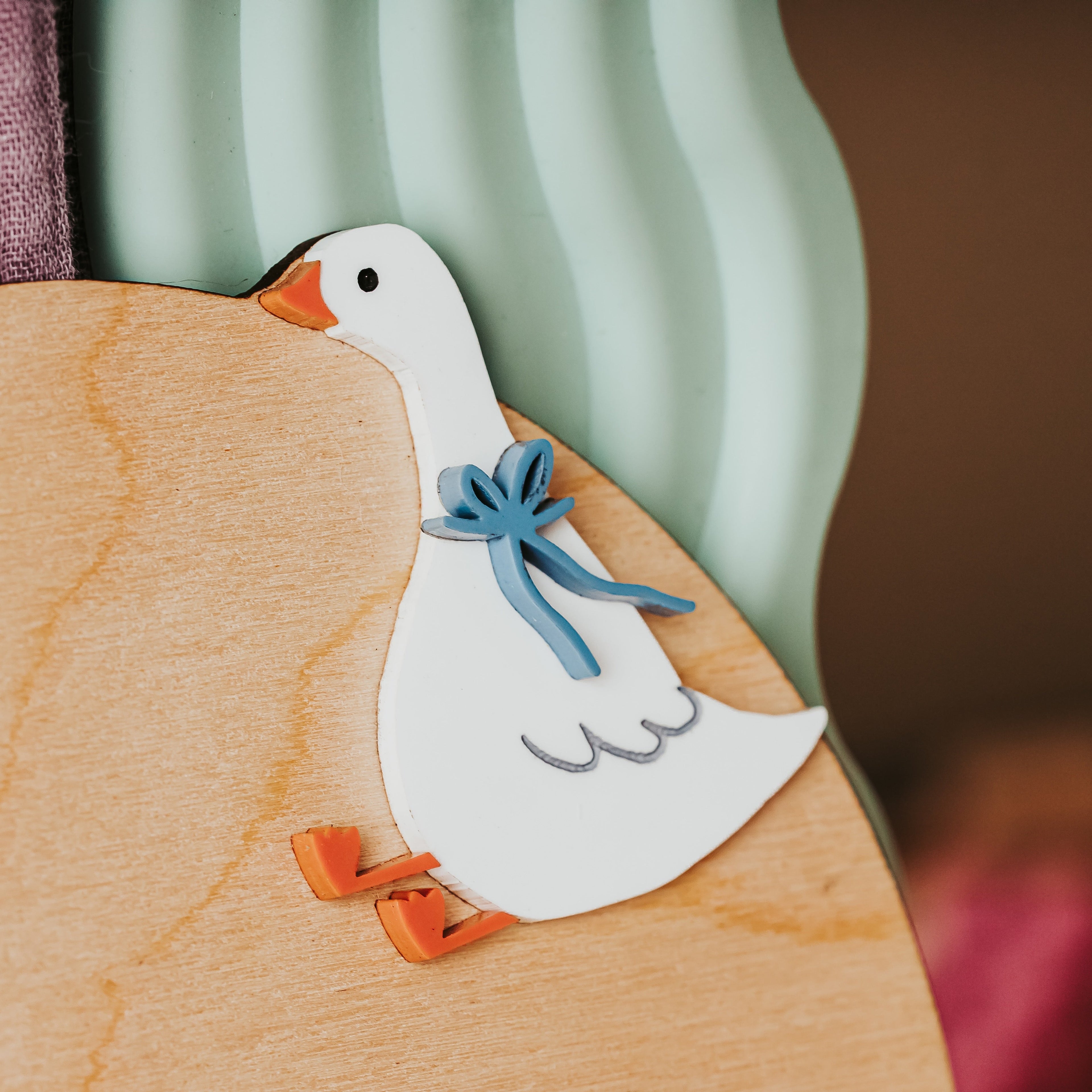 A small white goose-shaped decorative item with orange feet and beak, and a blue bow around its neck, resting on a light wood surface.