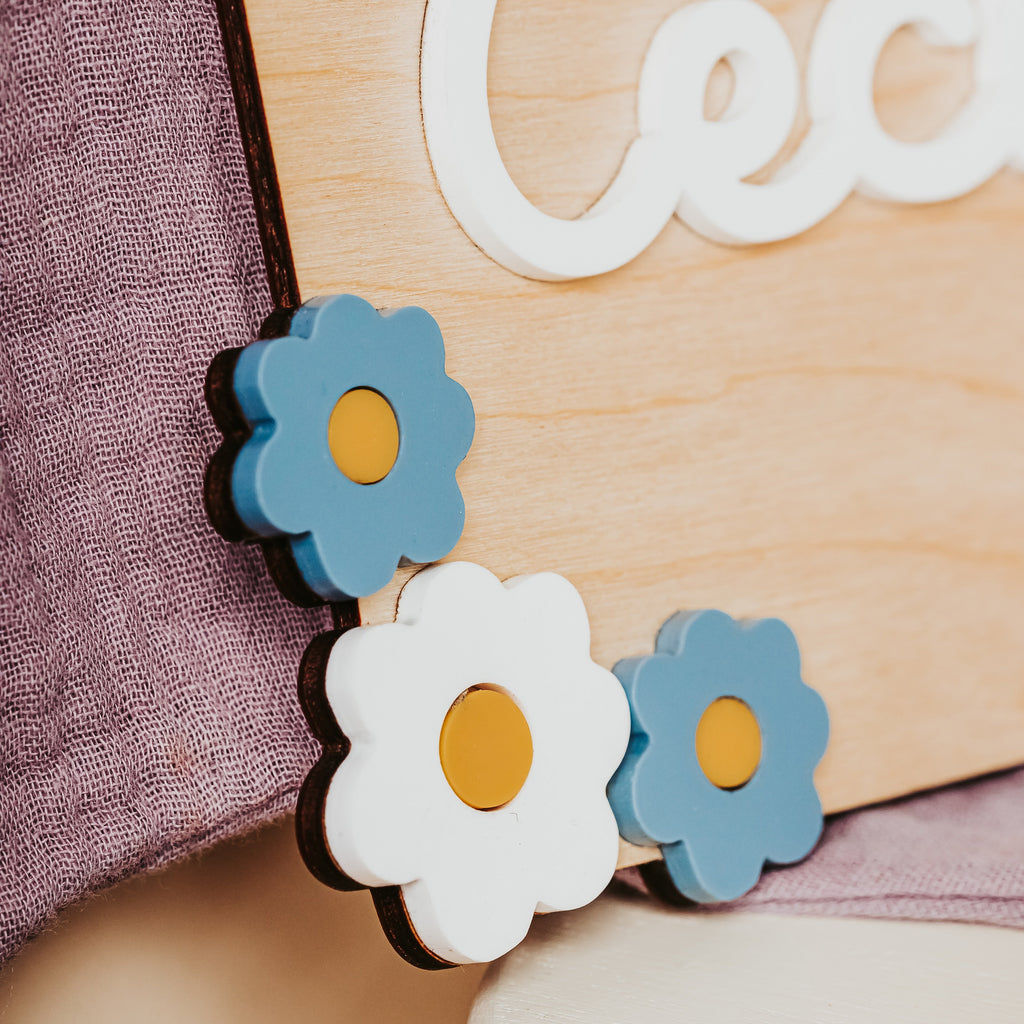 Three wooden flower-shaped baby name sign with blue petals, white petals, and yellow centers, shown resting on a light wood sign with a purple textured fabric background.